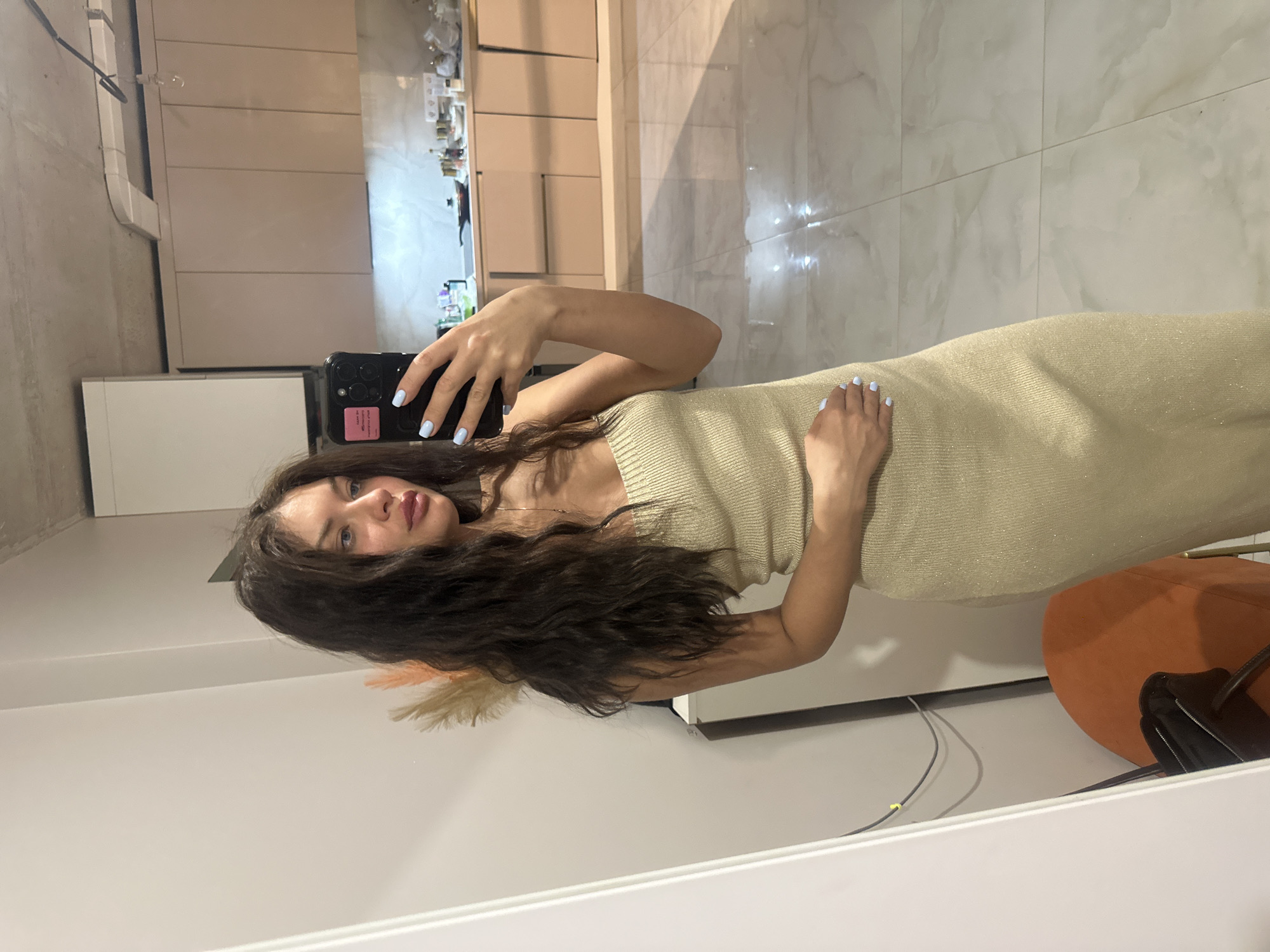 A woman with long dark hair is taking a mirror selfie in what appears to be an apartment or home. She is wearing a beige or light gold dress. Her nails are painted light blue. The background includes kitchen cabinets, a marble-tiled floor, a white wall, and part of an orange chair. The location is Unknown, Unknown.