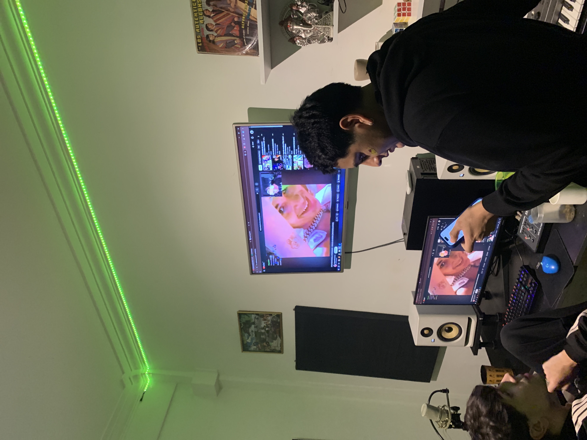 Two individuals are present in what appears to be a recording studio or music production workspace. One person, wearing a black hoodie, is standing and interacting with a computer monitor. Their hand is visible, touching the screen. The monitor displays a video or image of a person wearing jewelry. A second monitor is mounted on the wall, also displaying the same content. Additional studio equipment is visible, including speakers, a microphone, a keyboard, and other electronic devices. The room has a green LED strip along the ceiling. The top-left corner of the wall has a shelf with vinyl records and figurines. The location is unclear, and there are no clear signs to indicate Buenos Aires, Argentina.