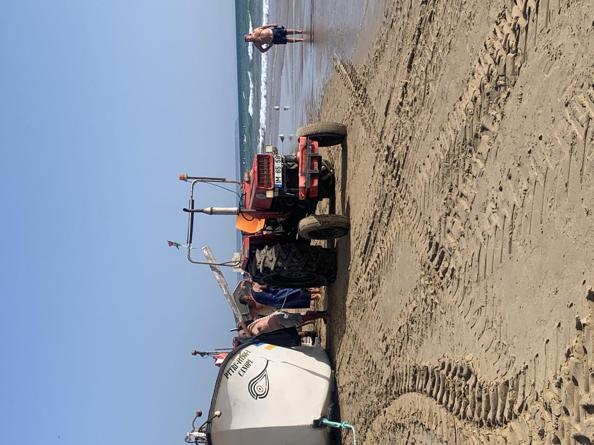 This image depicts a sunny day on the beach in Costa da Caparica, Portugal. In the foreground, a large, red tractor with the license plate "IM 65 58" is parked on the sand. Tire tracks are visible in the sand, indicating recent movement of heavy machinery. To the left of the tractor, a white fishing boat rests on the shore, with the inscription "PTTRF-44966-L CANOPE" on its side. Several individuals, appearing to be men, are interacting with the boat, with some partially obscured from view. One man, wearing dark shorts and a cap, stands in the shallow water to the right of the tractor, facing away from the camera. The ocean stretches to the horizon, with gentle waves breaking on the shore. The sky is clear and blue, with a faint airplane trail visible in the upper left corner. The overall scene suggests a maritime activity, possibly involving the launching or retrieval of the fishing boat with the assistance of the tractor.