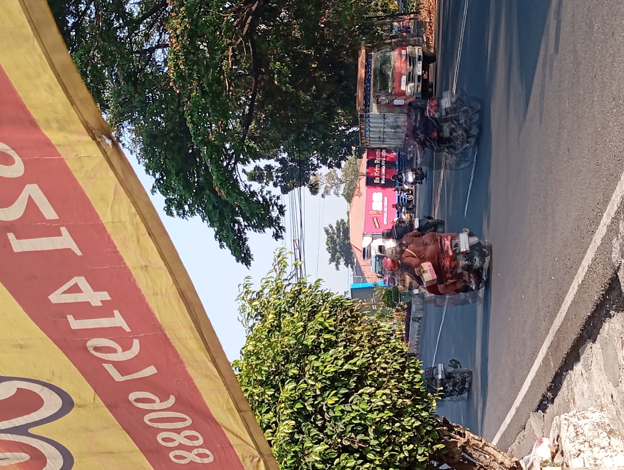 The image shows a street scene in Pandaan, Indonesia. In the foreground, a banner with red and yellow stripes stretches across the top of the frame, displaying white lettering that appears to read "880e ге14 ISO". The street is paved with asphalt and a white line marks the edge of the road. Several motorcycles are visible, some with riders, moving along the road. One motorcycle in the center has motion blur, suggesting it is in motion. On the right side of the road, a red truck with a white cargo container is parked. Further down the road, a pink building with large white text and graphics is visible. Trees with lush green foliage line the sides of the road. The sky is clear and blue, indicating a sunny day. The overall impression is of a busy street in a tropical or subtropical urban environment.