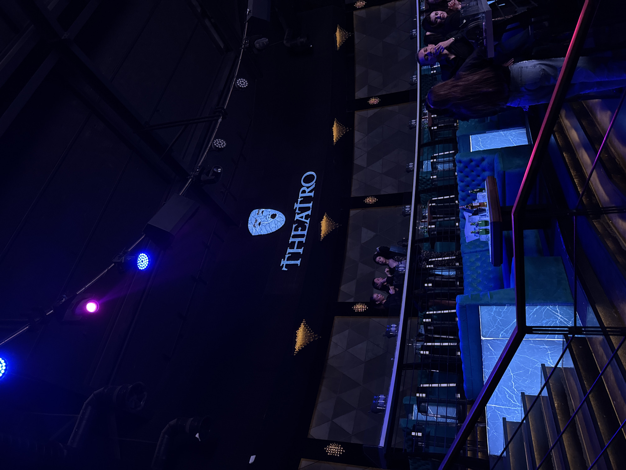 The image shows an interior shot of Teatro, located in Varna, Bulgaria. The "TEATRO" logo, accompanied by a mask-like symbol, is prominently displayed on a large black wall. A multi-tiered seating area is visible to the right, occupied by several people. The seating features plush blue velvet-like material and is illuminated with blue light. The environment seems to be a stylish nightclub or lounge, with a mix of patrons enjoying the atmosphere. A staircase with metal railings is visible on the right side of the image. Lighting fixtures and structures are visible on the ceiling.