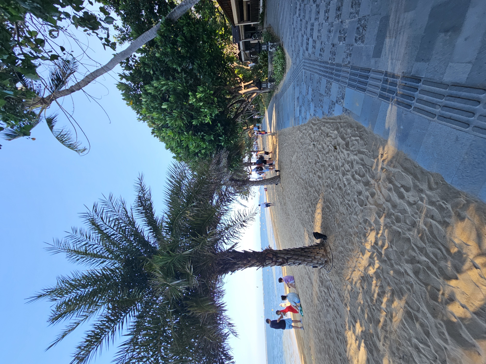 This is a vertical, eye-level shot of a beach pathway and sandy shore at Sanur Beach, Denpasar, Indonesia. In the foreground, a sandy beach is illuminated by dappled sunlight casting shadows from palm trees. Several people are seated on the sand near a palm tree, including a man in a blue shirt and shorts, a woman in a red dress, and two others in casual attire. Further down the beach, other individuals are walking and strolling. To the right of the sand, a paved walkway made of grey stone slabs with embedded tactile paving runs parallel to the beach. Lush green trees line the pathway, and beyond them, a low-rise building with a dark roof is visible. The ocean stretches to the horizon under a clear blue sky, with a few small boats and a distant structure visible on the water.