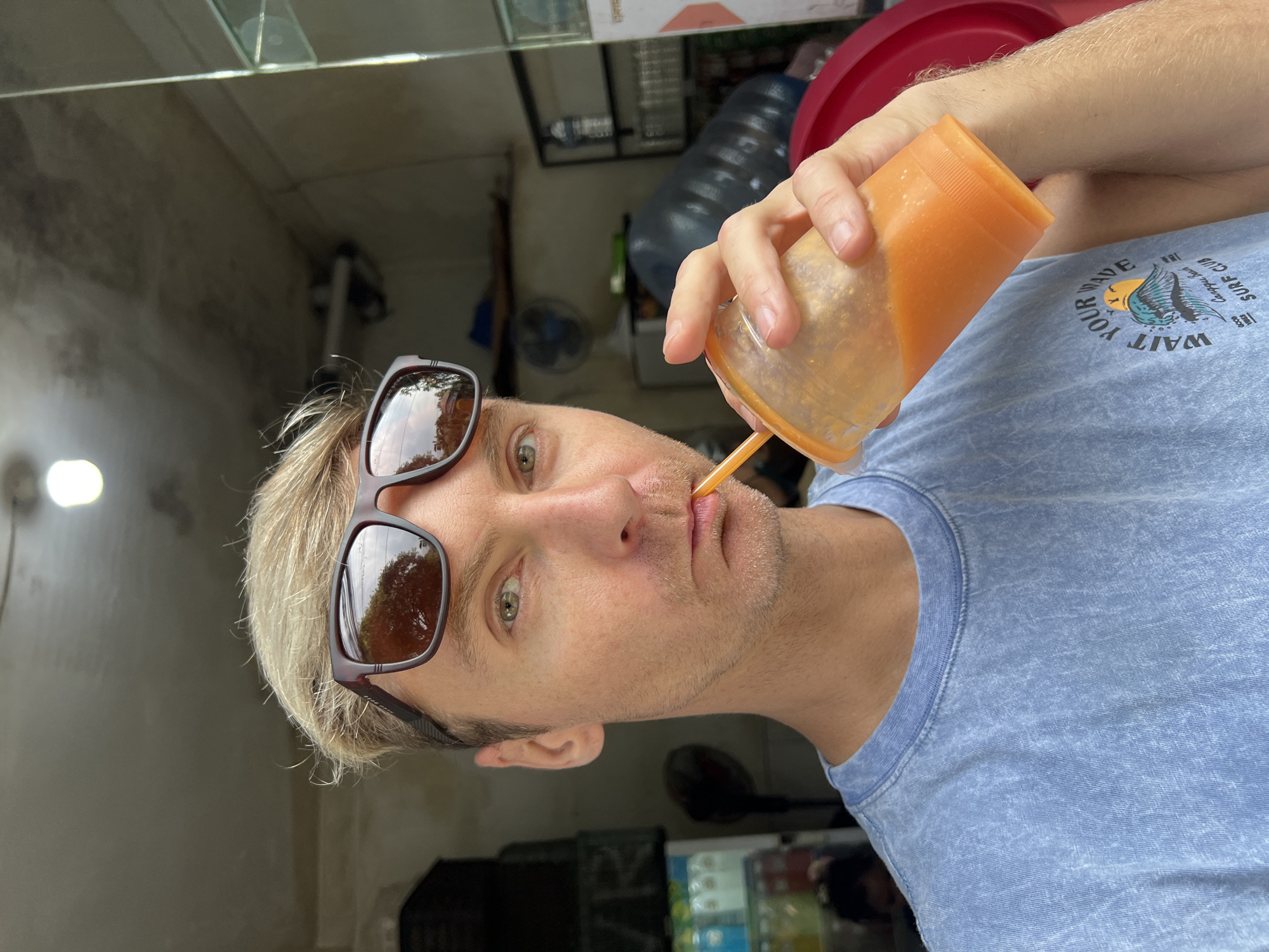 A fair-skinned man with blond hair, stubble, and sunglasses is drinking from an orange plastic cup with a straw. He is wearing a blue t-shirt with a graphic design on it. The background is a partially visible interior space, possibly a small shop or eatery in Jimbaran, Indonesia. A fan and plastic water bottles are visible in the background. A display case can also be seen near the bottom of the image.
