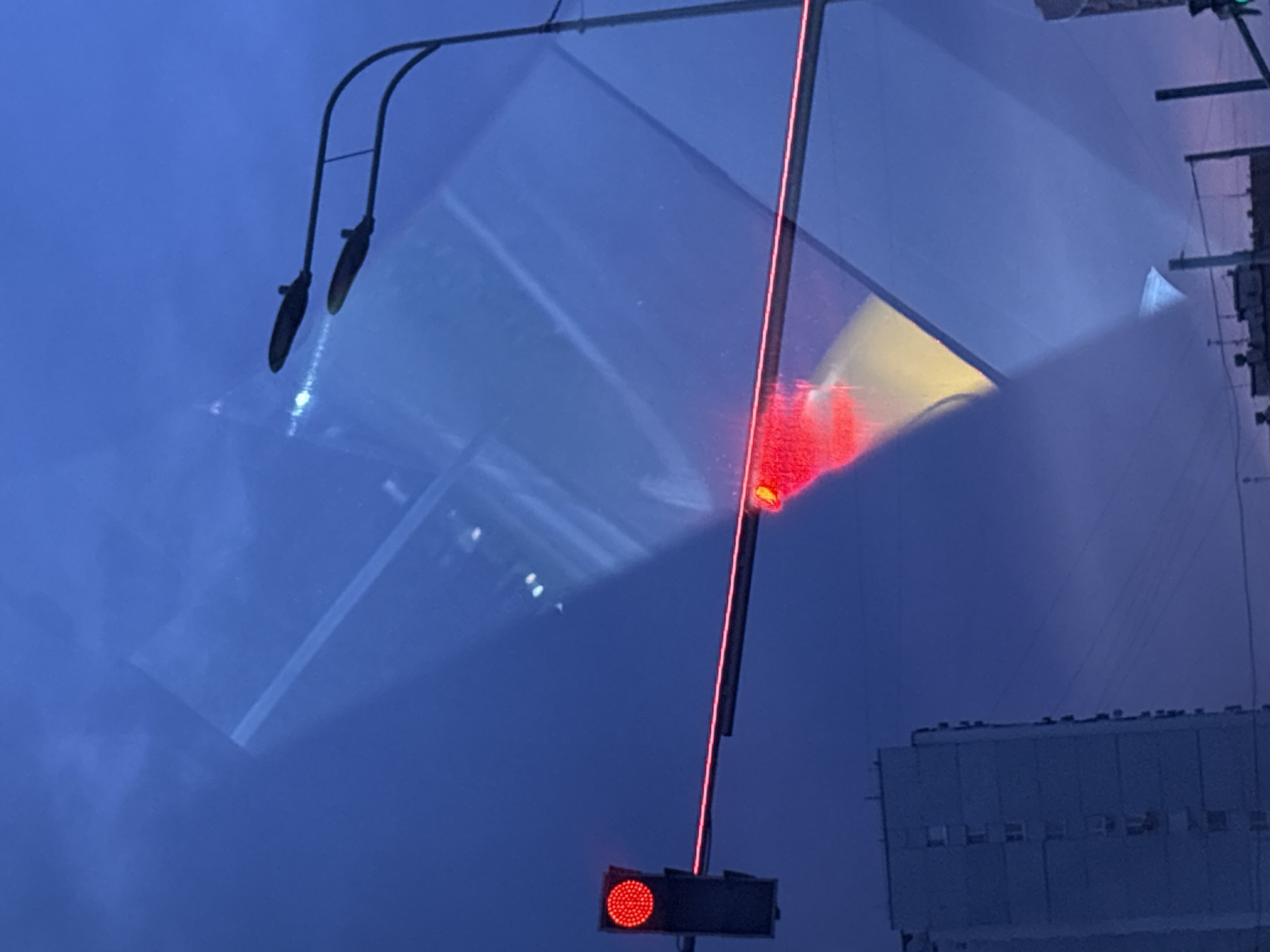 The image captures an urban scene at dusk or night. The primary subject is a traffic signal, displaying a red light. This signal is positioned in the foreground, partially obscured by reflections. Above the traffic signal, a thin red line of light extends upwards, likely from a decorative element or another light source. In the background, a multi-story building with a row of windows is visible. The sky is a deep blue, suggesting twilight or nighttime.
The image is dominated by reflections and light. There are prominent, distorted reflections of streetlights or architectural elements on a smooth, possibly wet or glass surface. These reflections create geometric patterns and streaks of light across the scene, including a yellow and red glow. The overall impression is one of an urban environment, possibly a street or plaza, with artificial lighting playing a significant role in the visual composition. The weather appears to be clear, given the sharp reflections. No people are visible in the image. There is no discernible text.