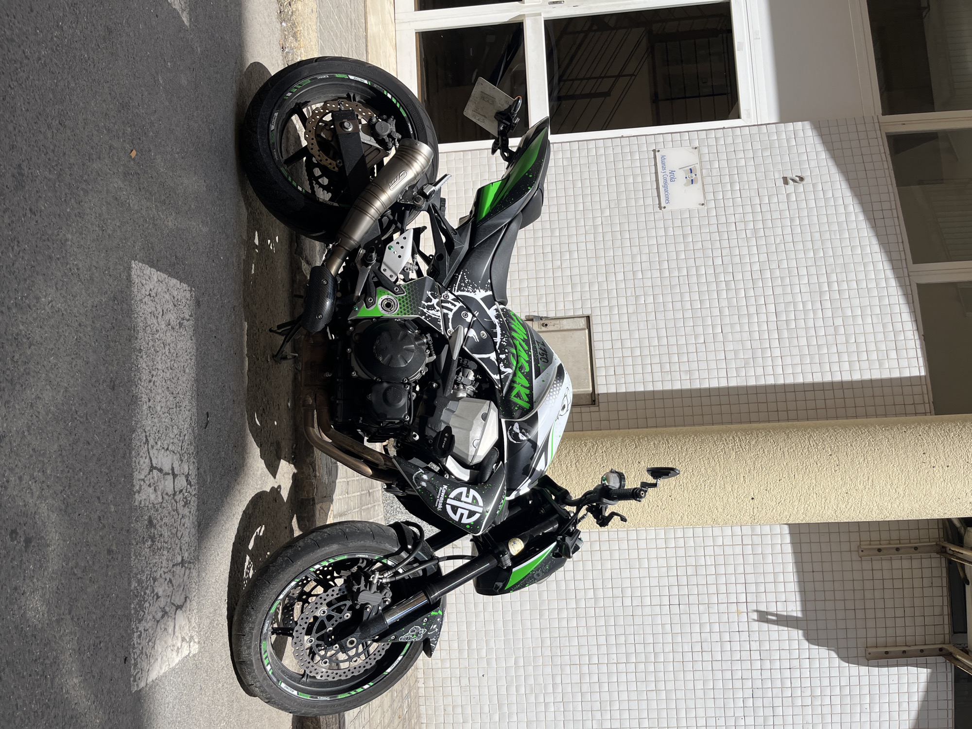 The image shows a black and green Kawasaki Z900 motorcycle parked on a street in Tarragona, Spain. The motorcycle is positioned diagonally in the frame, facing towards the right. It features a custom graphic design with white splatters and green accents. The building behind the motorcycle has a facade covered in white square tiles. A cream-colored cylindrical pillar is located to the right of the motorcycle. Above the motorcycle, a white sign is visible on the tiled wall, with text in Spanish that appears to be "Arola Aduanas y Consignaciones". A large number "2" is also visible on the tiled wall, above the sign. The street in front of the motorcycle has a white pedestrian crossing marking. The lighting suggests a sunny day, with strong shadows cast by the pillar and the motorcycle onto the street and pavement. There are no people visible in the image.