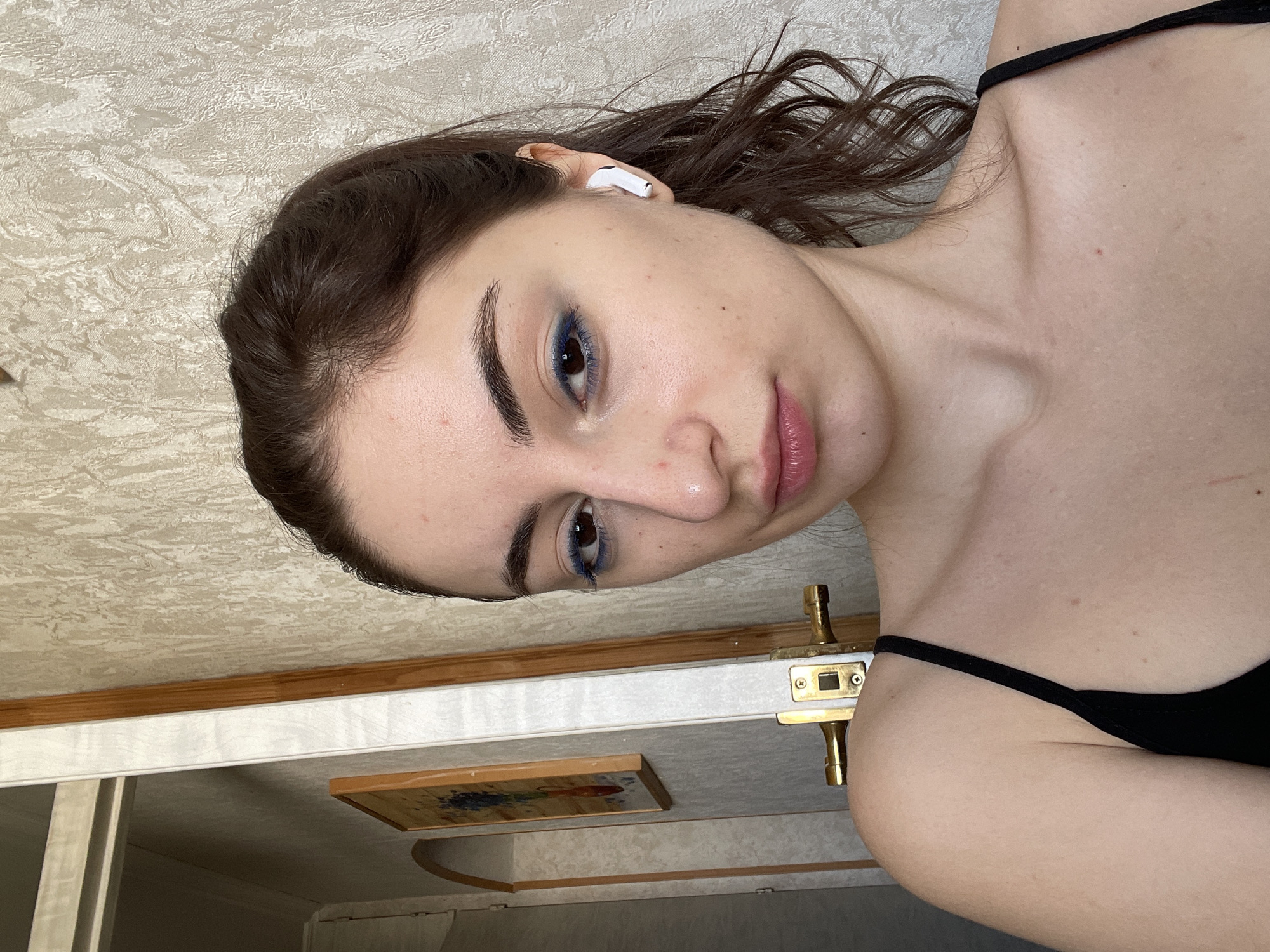 A young woman with dark hair is positioned in the frame, wearing a black strappy top and a white wireless earbud in her left ear. She is looking at the camera, her face showing light makeup with noticeable blue eyeliner.
The background appears to be an interior scene. A decorative wall or ceiling panel is visible with a textured pattern and light color. An ornate door handle and lock are positioned nearby, and a framed picture is partially visible on a wall beneath it.
There is no verifiable information in the image to ascertain the location of the scene as Moscow, Russia.