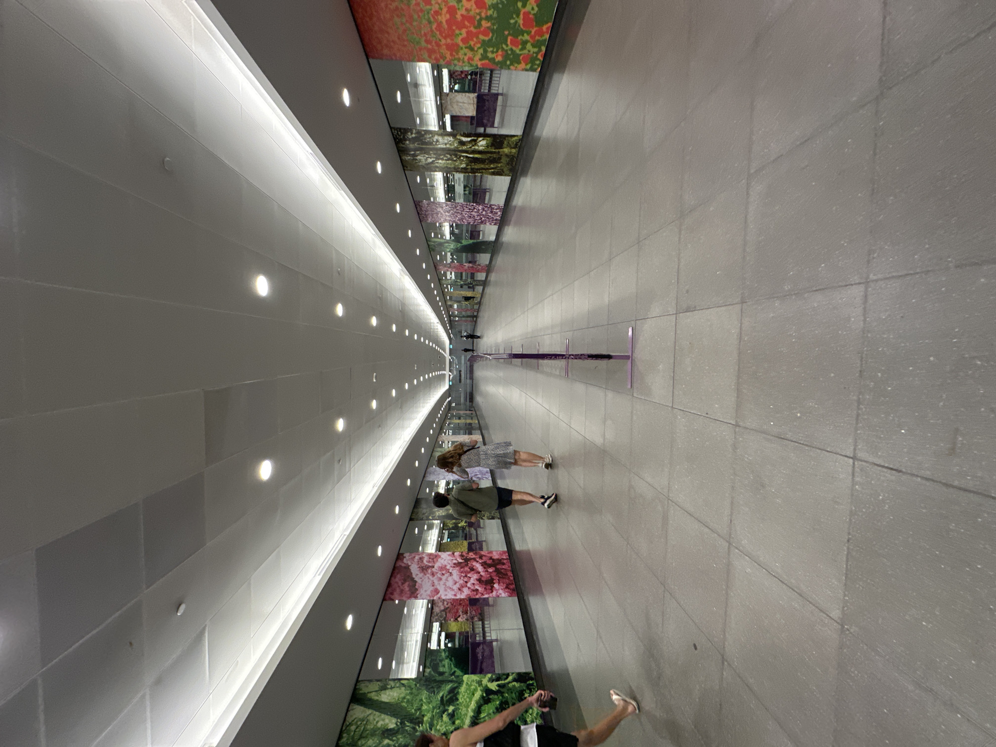 The image shows a long, wide, indoor pedestrian walkway with a high, illuminated ceiling. The walls are lined with large, colorful photographic murals of natural scenes, interspersed with reflective panels. Several people are visible walking in the walkway. In the foreground, on the left, a person wearing shorts and a tank top walks away from the camera. Further down the walkway, two people, a man and a woman, walk away from the camera. In the distance, other figures are faintly visible. Several purple metal stanchions with clear panels are positioned along the walkway. The floor is tiled with large, grey square tiles. The overall impression is of a modern, well-lit public transit station or airport concourse. The location is identified as Singapore, Singapore.