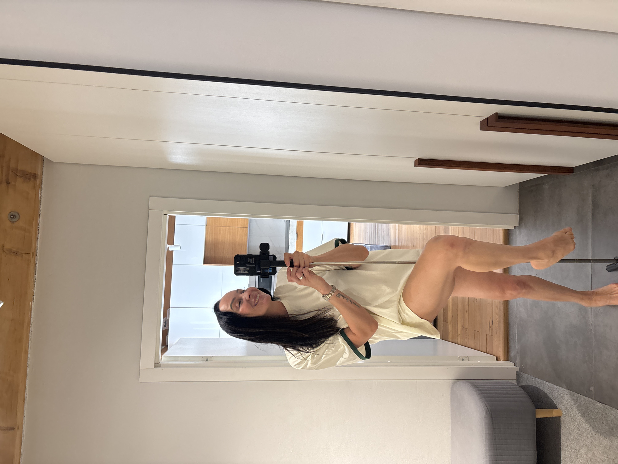 A female individual is depicted in an indoor setting in Chaikovsky, Russia, capturing a self-portrait using a black smartphone mounted on a metallic selfie stick/tripod. She has long dark hair, a visible tattoo on her right forearm, a watch on her left wrist, and a ring on her left ring finger. She is wearing an oversized, light-cream t-shirt with dark green trim on the sleeves, is barefoot, and presents a smiling expression towards her reflection. Her right leg is bent at the knee, with the foot resting on the selfie stick's base. She stands on a dark grey tiled floor, reflected in a full-length mirror, which forms part of a white built-in wardrobe system featuring dark wooden vertical handles. To the left, a grey upholstered ottoman with wooden legs is positioned. A white-framed doorway behind the individual opens to an adjacent room with light wooden plank flooring, revealing modern white and wooden kitchen-style cabinetry and integrated appliances. Exposed wooden beams are visible on the ceiling above.
