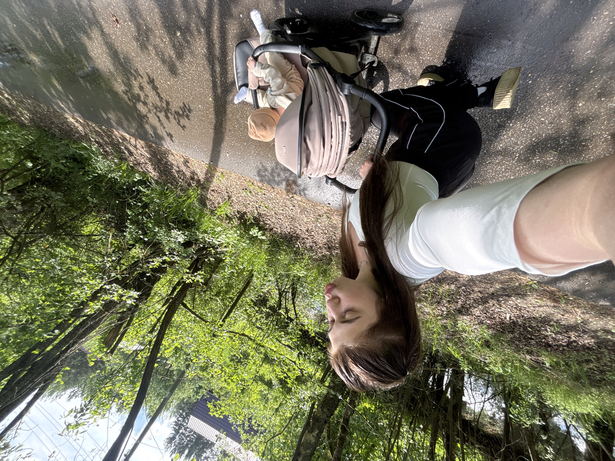 A young woman is taking a selfie while pushing a baby stroller on a paved path. The path is surrounded by trees and vegetation, suggesting a park or green area. The baby in the stroller is wearing a hat and is bundled up in a blanket. The woman is wearing a white top and black pants. The location could be in Мещёры, Russia, given the prompt's instruction.