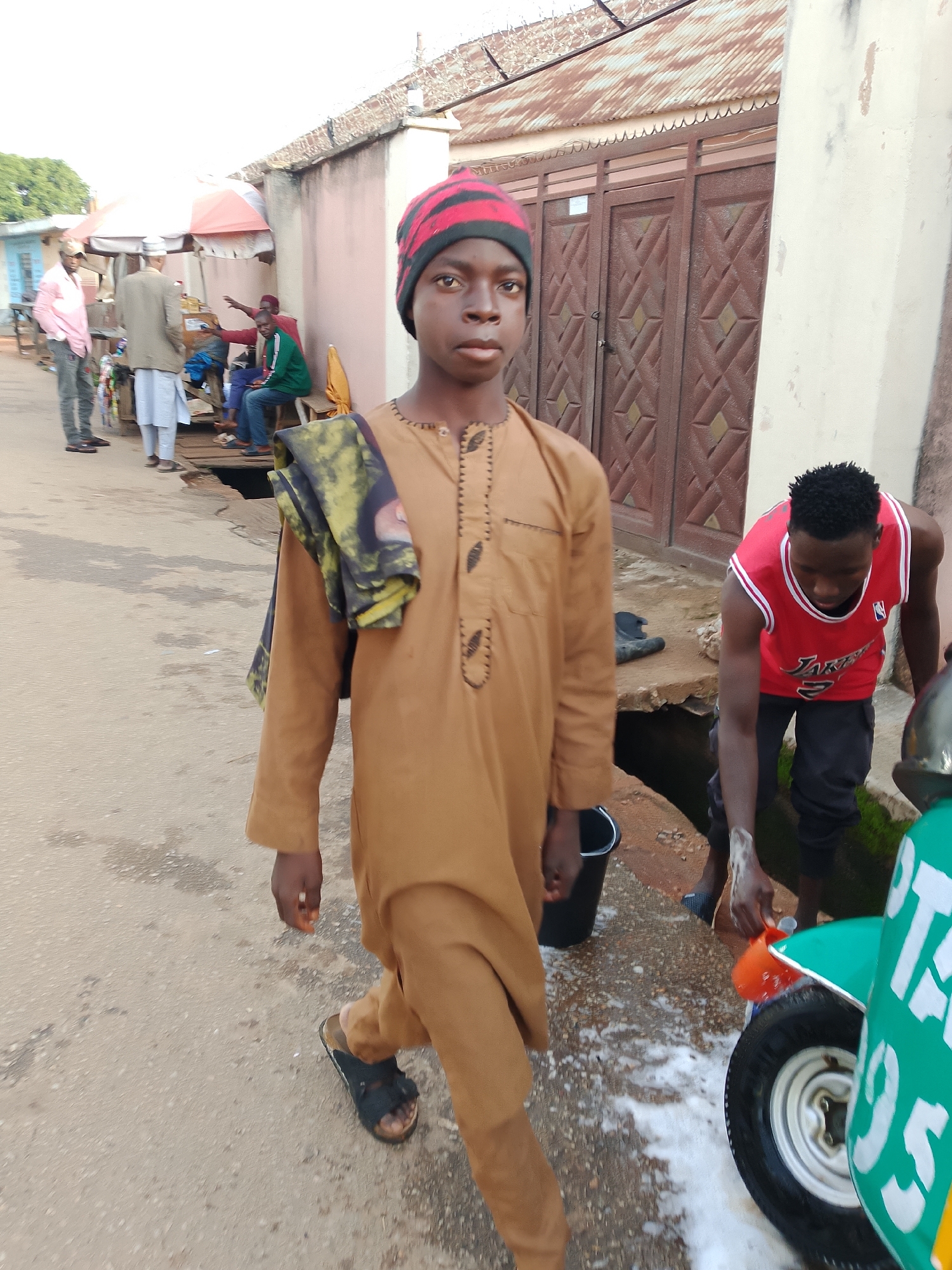 In Jos, Nigeria, on a sunlit day, a young person stands in the foreground, wearing a tan kaftan and a red and black striped beanie. They carry a patterned cloth over their shoulder. Behind them, several individuals are gathered near a stall with an umbrella, suggesting a market or roadside vendor area. One person in a red basketball jersey is washing a green and white vehicle, possibly a commercial tricycle, with soapy water. The environment appears to be a somewhat unpaved street with a drain running alongside. The buildings in the background have corrugated iron roofs and wooden doors. The scene depicts everyday street life with commercial activity and personal hygiene taking place outdoors.