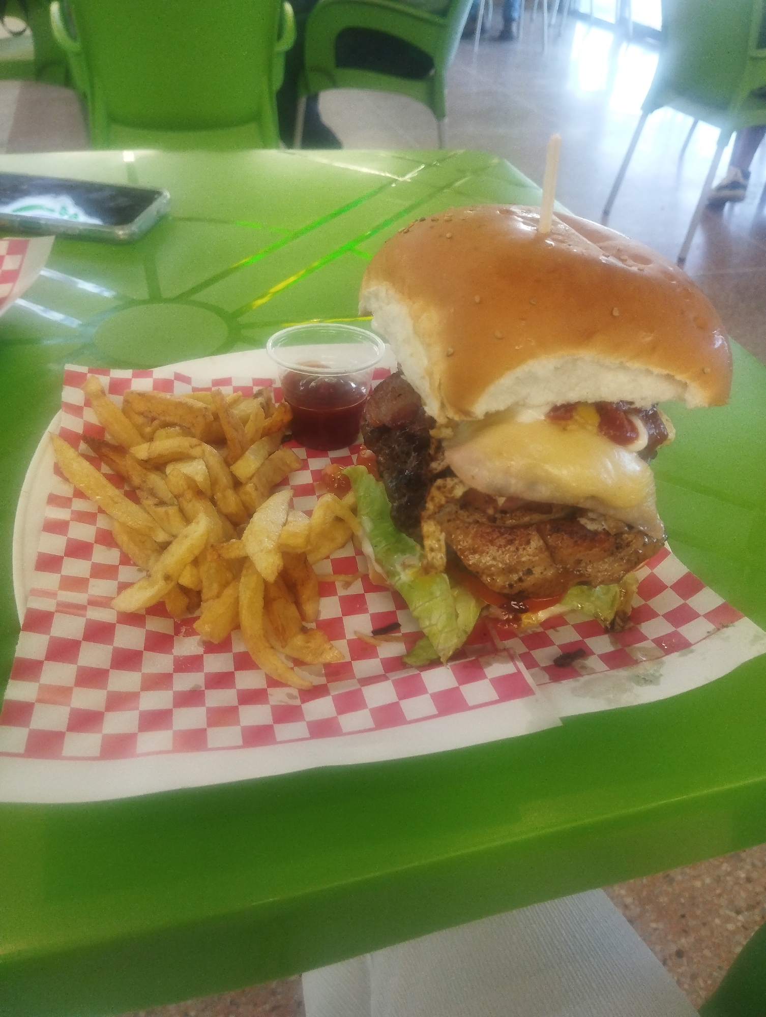 The image shows a large hamburger with fries and a small container of sauce on a plate lined with red and white checkered paper. A toothpick is stuck in the top of the bun. The plate rests on a bright green table. A smartphone is visible on the table to the left of the plate. The scene appears to be set indoors, with green chairs and tables visible in the background. Based on the prompt, the location is a restaurant or eatery in the city of Porlamar, Venezuela.