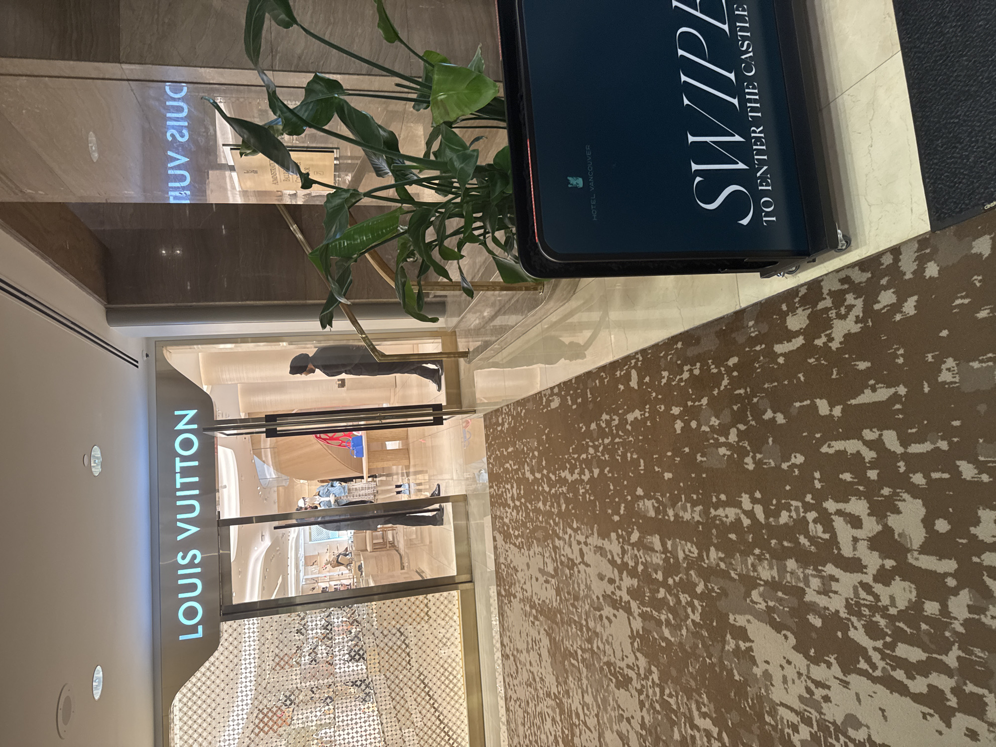 The image shows the exterior of a Louis Vuitton store. The entrance is visible with the words "LOUIS VUITTON" vertically aligned above the entrance. Inside the store, a person is seen walking. A plant and an advertising display for the Hotel Vancouver are in the foreground. The display reads, "SWIPE TO ENTER THE CASTLE" and shows the Hotel Vancouver logo. The interior design of the store features a checkered pattern near the entrance, suggesting a luxurious retail environment. The location is likely Vancouver, Canada, based on the Hotel Vancouver advertisement.