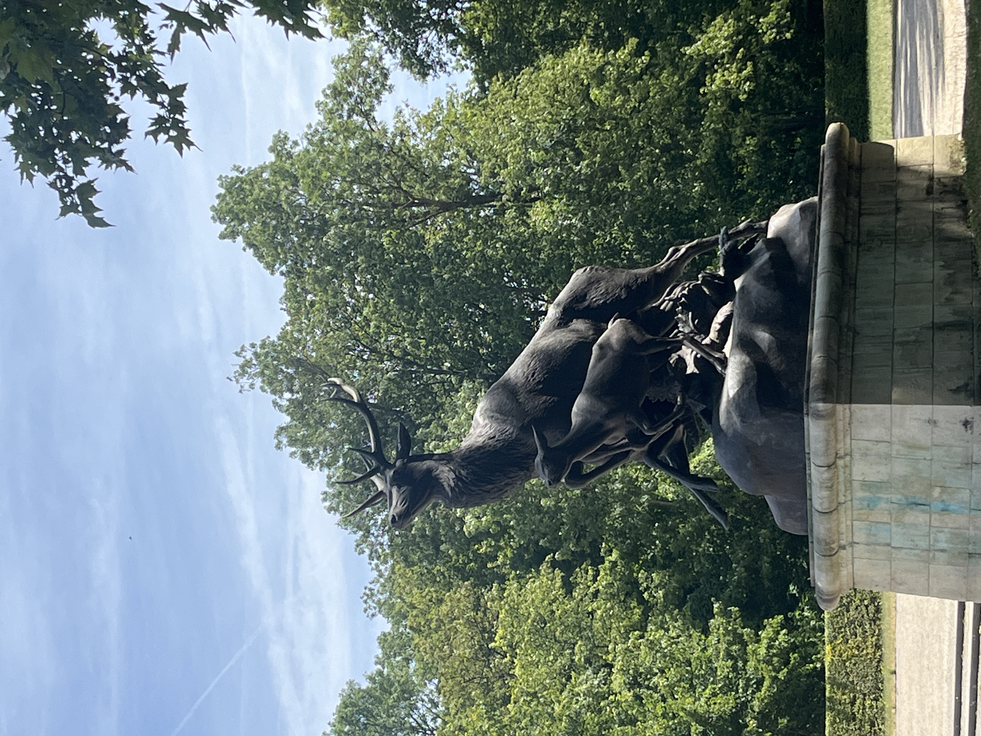 A bronze sculpture of a stag and two fawns stands on a stone pedestal. The stag, depicted in mid-leap, has its head raised and antlers spread. The fawns are positioned below and to the side of the stag, also in dynamic poses. The sculpture is situated in a park-like setting with lush green trees in the background. The sky above is blue with some scattered clouds and a contrail from an aircraft. The sculpture is located in the Parc de Sceaux, in Antony, France.