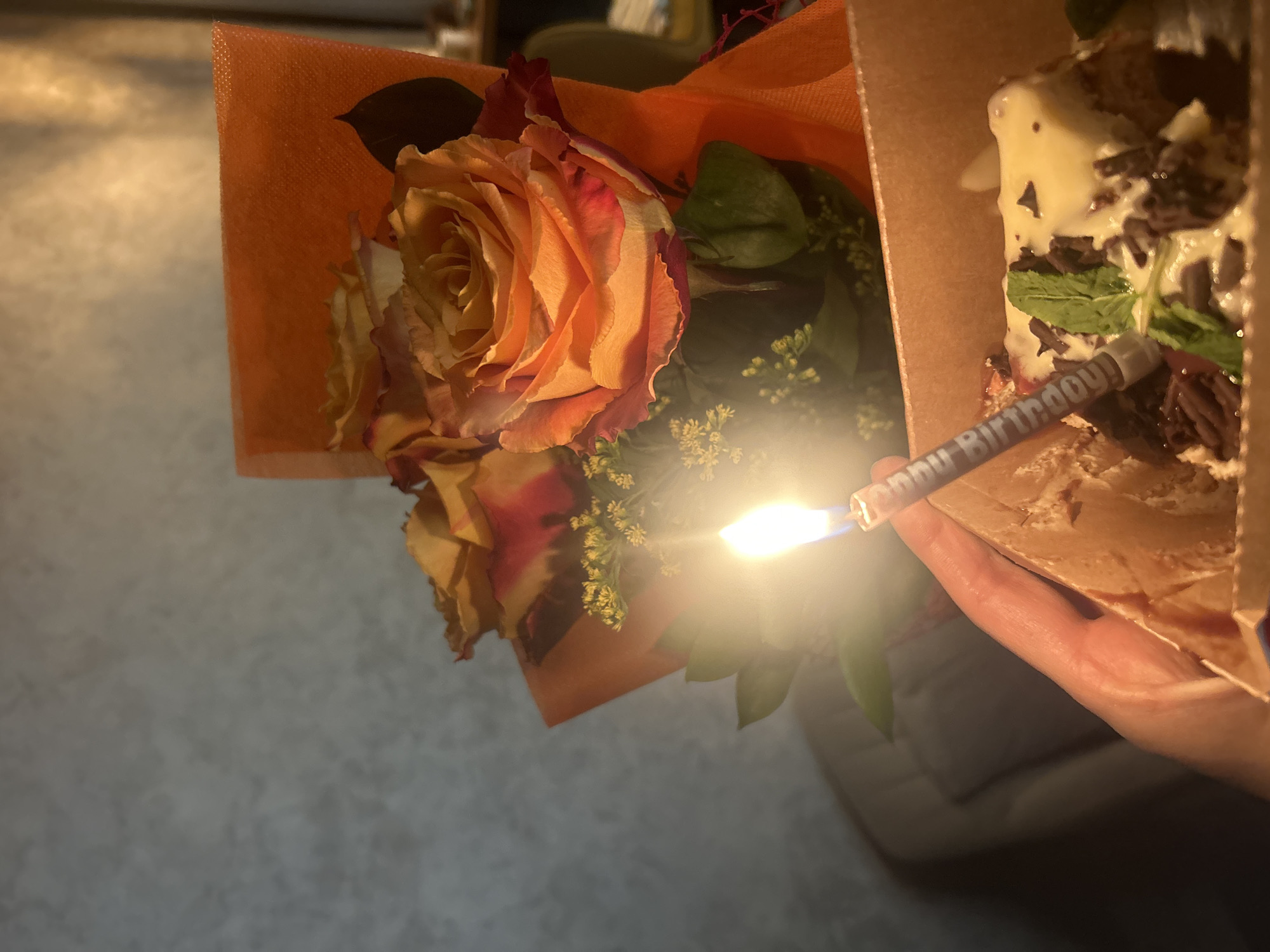 The image shows a close-up, angled view of a bouquet of flowers next to a slice of cake with a lit "Happy Birthday" candle. The flowers include a prominent peach and red rose. The cake appears to be topped with cream and chocolate shavings, with mint leaves as garnish. A hand is holding the box containing the cake. The background is blurred and nondescript, making it impossible to determine if it's in Sofia, Bulgaria or any other location. There are no people visible in full.