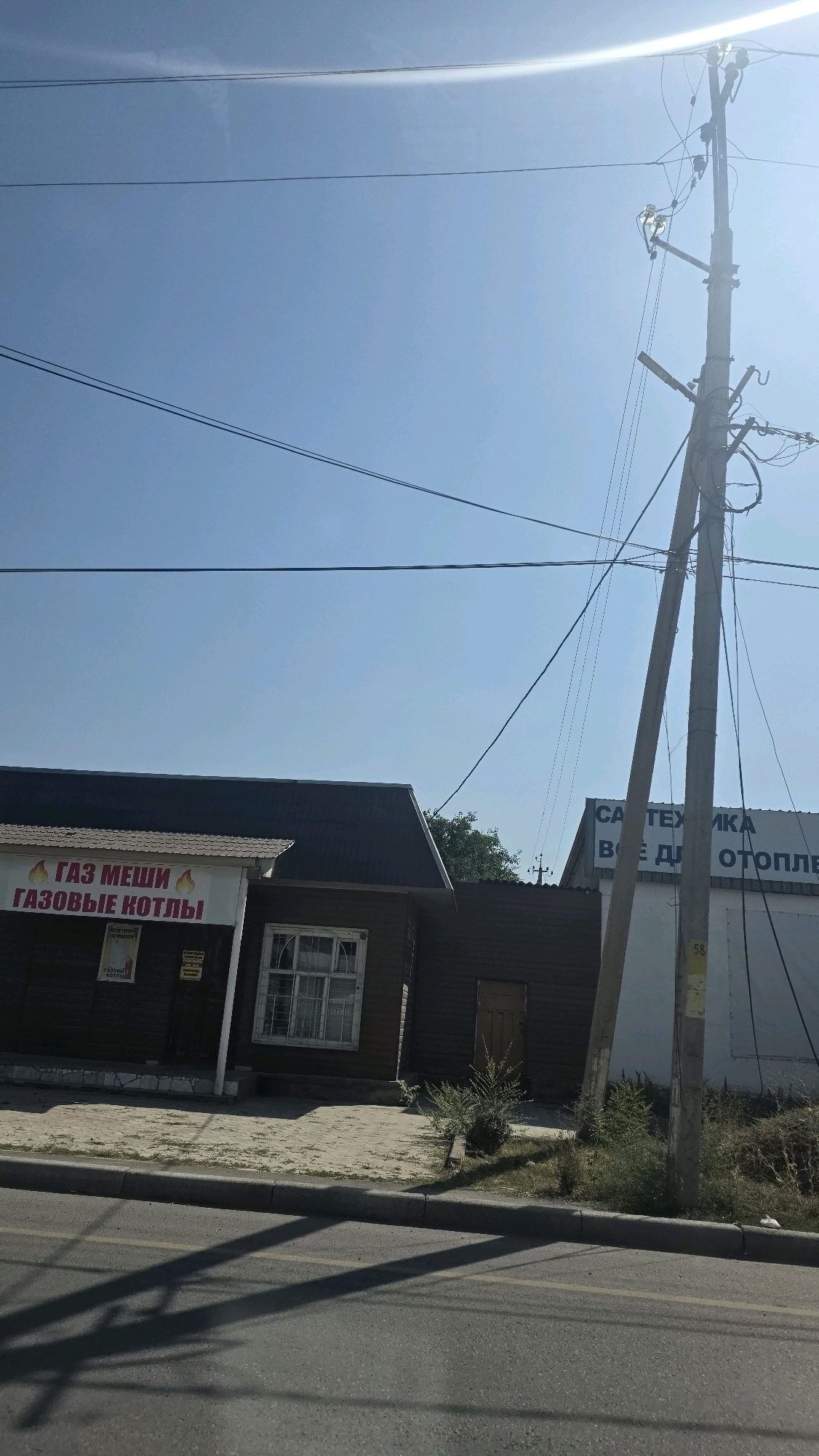An outdoor, eye-level, medium shot captures a sun-drenched street scene in Tokmok City, Kyrgyzstan. The sky is a clear, bright blue with the sun emitting a strong glare in the upper right corner. Power lines crisscross the sky, supported by a leaning utility pole on the right side of the frame.
In the foreground, a paved road curves slightly, with a concrete curb separating it from a tiled sidewalk. Shadows of power lines stretch across the road. Along the sidewalk are two buildings. The building on the left is a single-story structure with a dark, sloped roof. A sign with red lettering reads "ГАЗ МЕШИ ГАЗОВЫЕ КОТЛЫ" (Gas stoves, gas boilers). Adjacent to this sign are smaller posters.
To the right of this building is a darker, wooden building with a single window featuring a grate and a small, brown wooden door. A patch of sparse green bushes grows in front of this building. Further to the right, a large, white structure with dark lettering partially visible reads "СА ТЕХНИКА ВСЁ ДЛЯ ОТОПЛЕ" (Sanitary technology, everything for heating). A yellow number "58" is visible on the utility pole. The overall impression is of a commercial area on a bright, clear day.