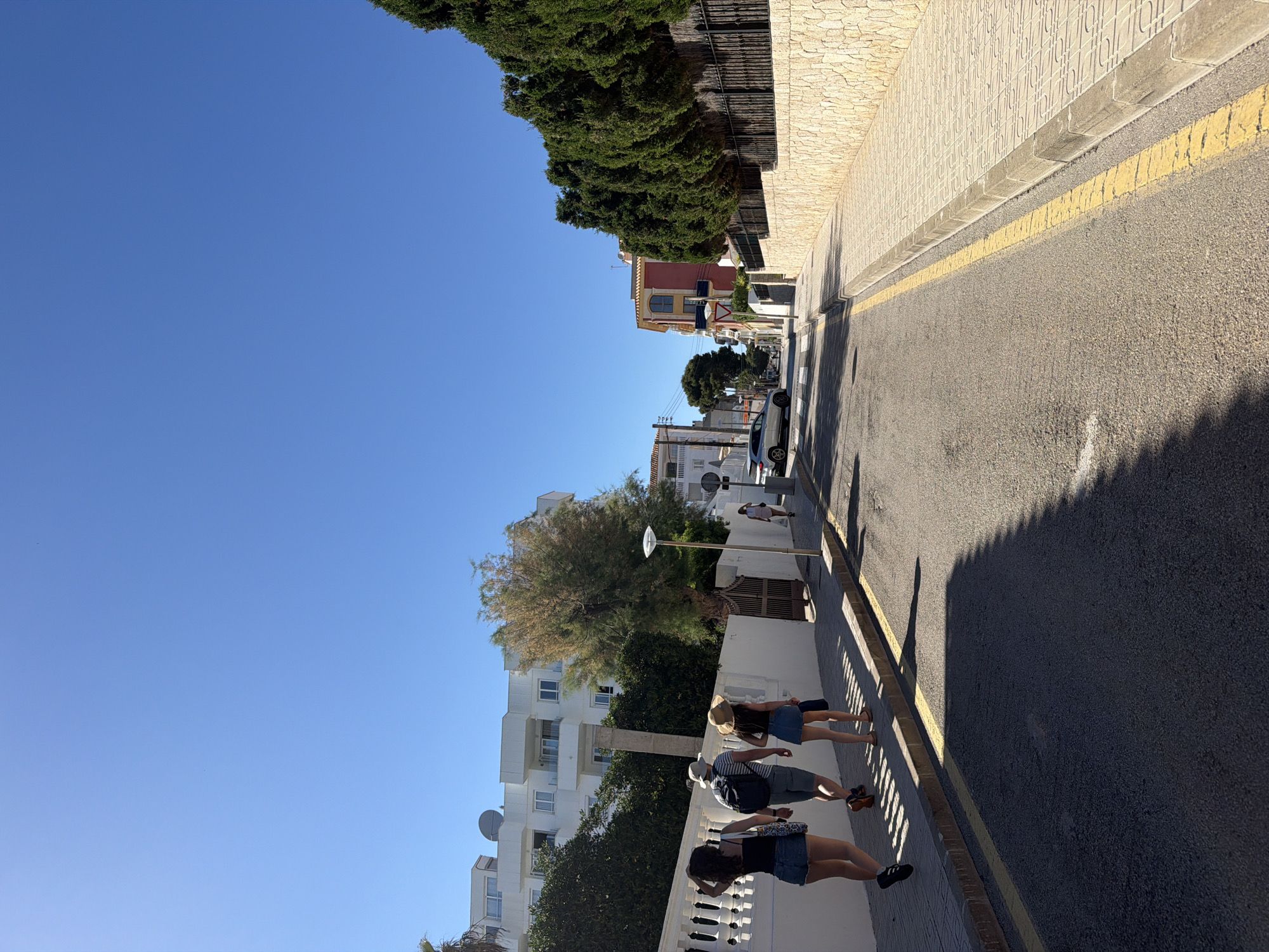 The image shows a street scene in Capdepera, Spain, on a sunny day. The clear blue sky contrasts with the varied architecture and vegetation lining the road. A group of three people are walking along the sidewalk, their shadows stretching onto the asphalt street marked with a yellow line. The buildings on one side are white with balconies and satellite dishes, while the other side features a stone wall topped with green trees. Further down the street, a mix of red and white buildings, parked cars, and a few additional pedestrians are visible. A tall street lamp stands next to a tree along the street.