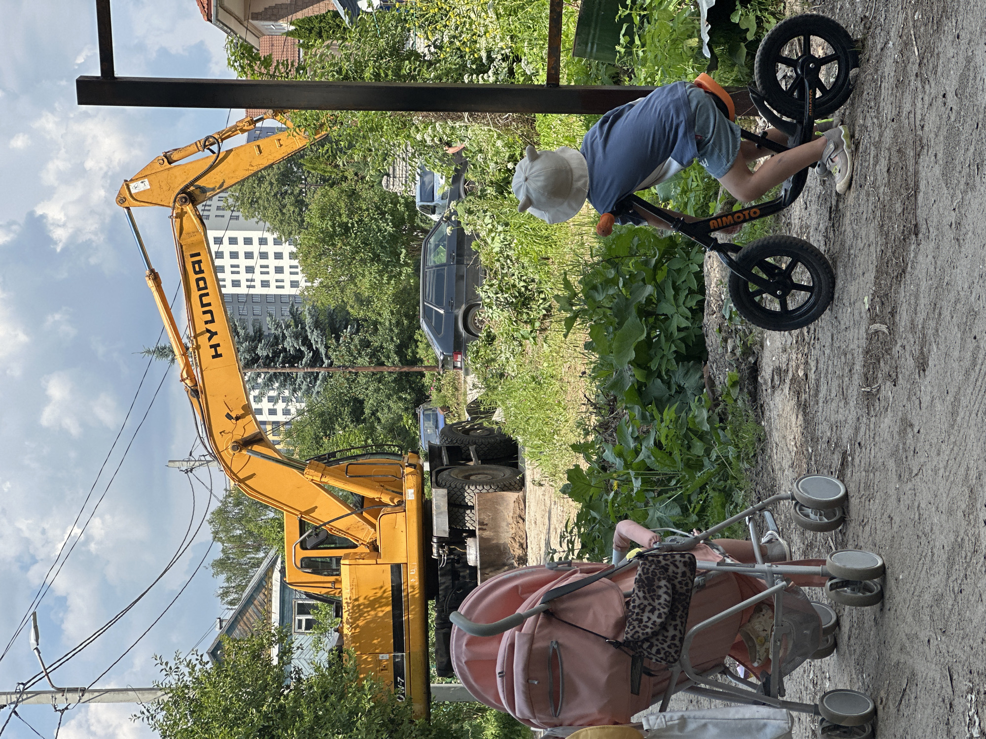 In Nizhny Novgorod, Russia, on a construction site, a yellow Hyundai excavator is parked near a residential area. In the foreground, a child rides a balance bike and a pink stroller sits beside them. The area is overgrown with greenery and there are several cars parked in the background. The sky is cloudy.