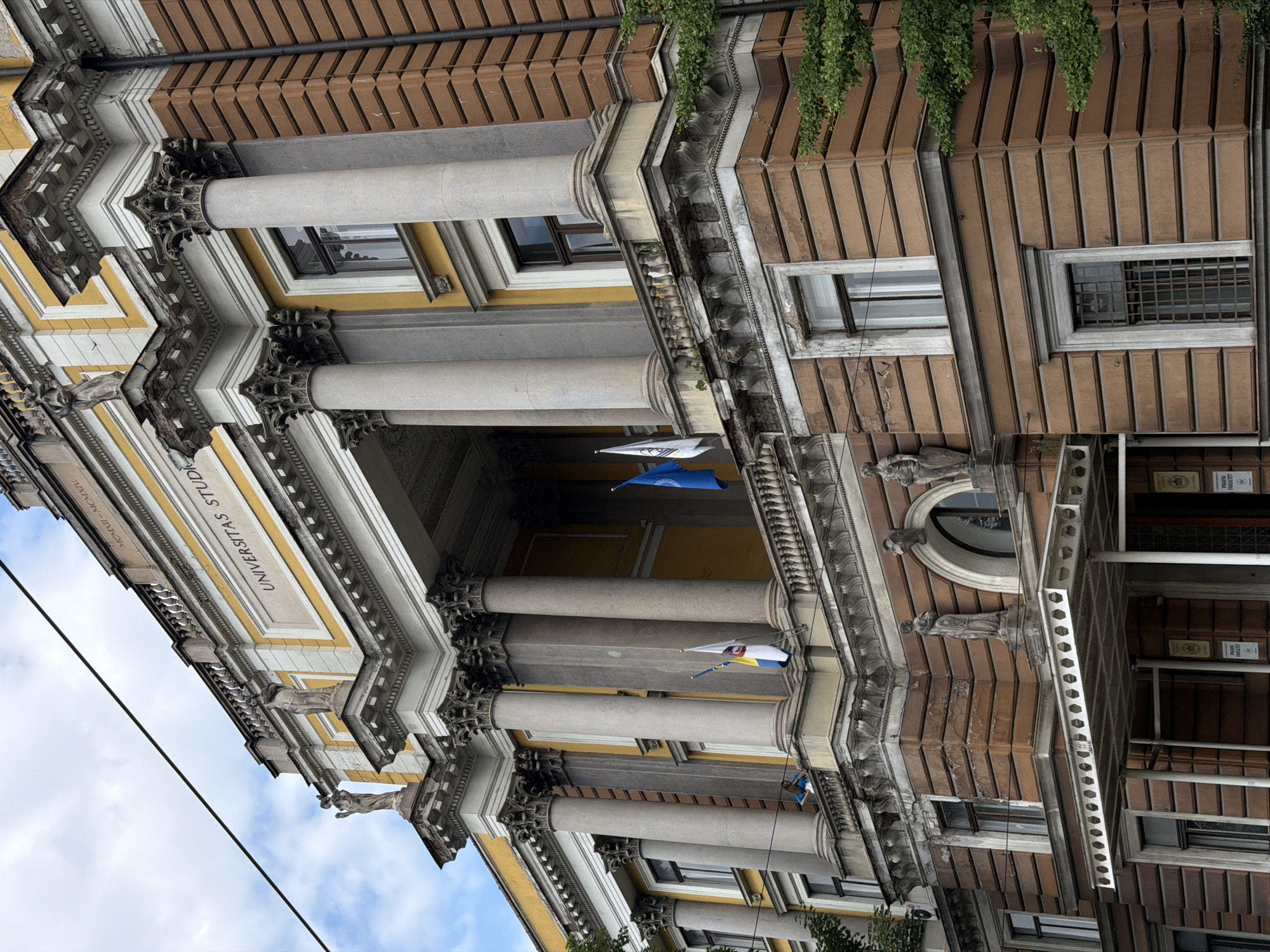 The image captures the imposing facade of the University of Sarajevo's Rectorate building, located in Sarajevo, Bosnia and Herzegovina. The neoclassical architectural style is evident, with grand columns, decorative statues, and ornate cornices. The building features a central portico with a prominent inscription that reads "UNIVERSITAS STUDIORUM." Two flags, one blue and one white with a coat of arms, are visible on a balcony. Below the main entrance, a smaller canopy shelters the doorway. A few people are visible on the balcony, and a single individual is standing on a lower ledge, looking out. The building is constructed with red brick and yellow stone accents. Lush green trees frame the right side of the building, and a power line crosses diagonally across the upper portion of the image. The sky is partly cloudy.