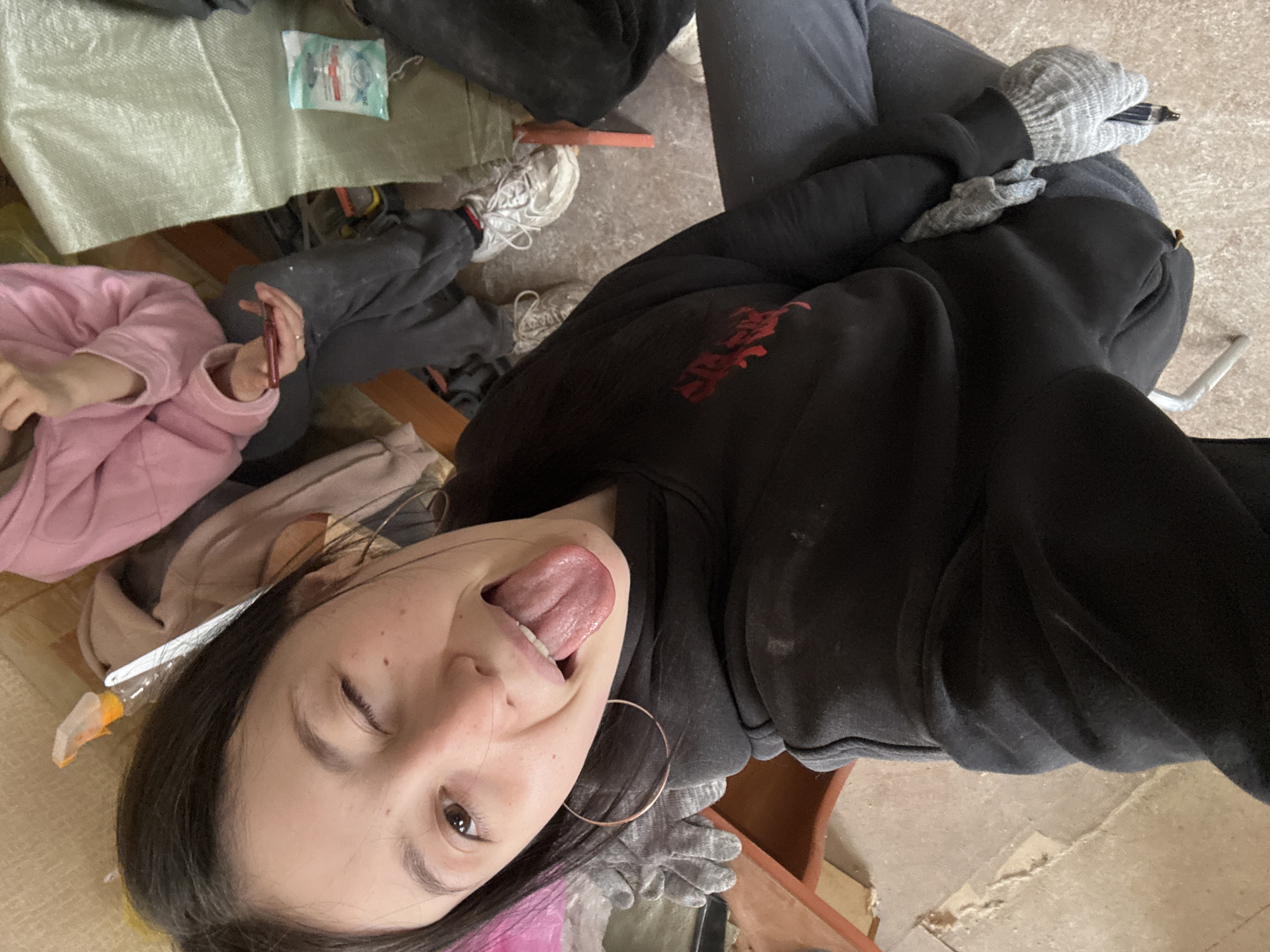 A selfie-style photograph shows a woman sticking out her tongue and winking. She has dark hair and wears large hoop earrings, a black hooded sweatshirt, and gray gloves. A portion of a pink garment is visible near her face. The background includes work clothing, a gray bag, white tennis shoes, and what appears to be a plastic container of wipes. The context suggests a construction site environment, possibly in the city of Фоки, Russia.