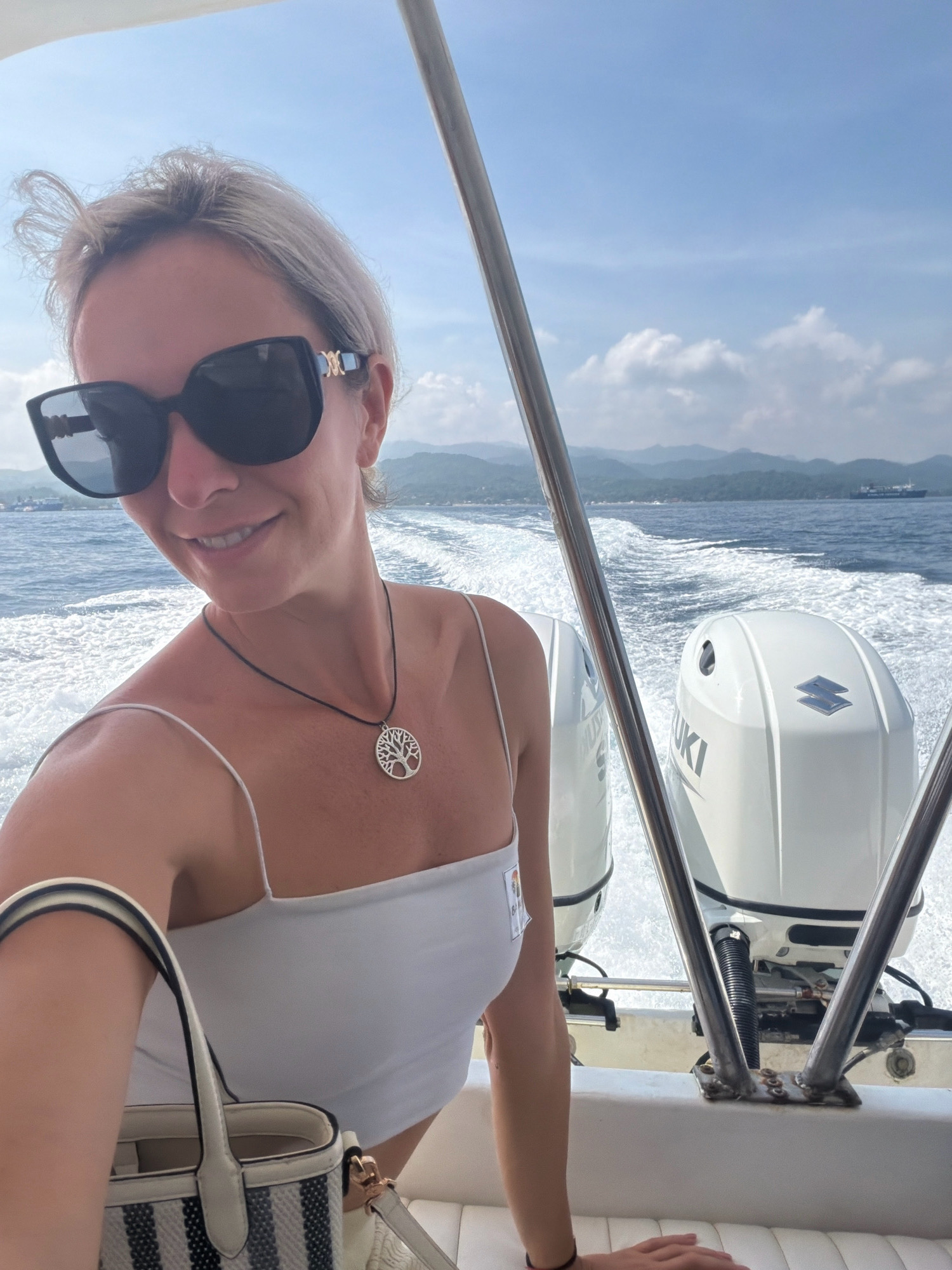 A woman with blonde hair and sunglasses is on a boat, taking a selfie. She is wearing a white tank top and a necklace with a tree of life pendant. She is holding a striped purse. In the background, two outboard motors are visible. The boat is moving through the water, creating a wake. The scenery suggests a coastal area with mountains in the distance, consistent with the location of Malay, Philippines.