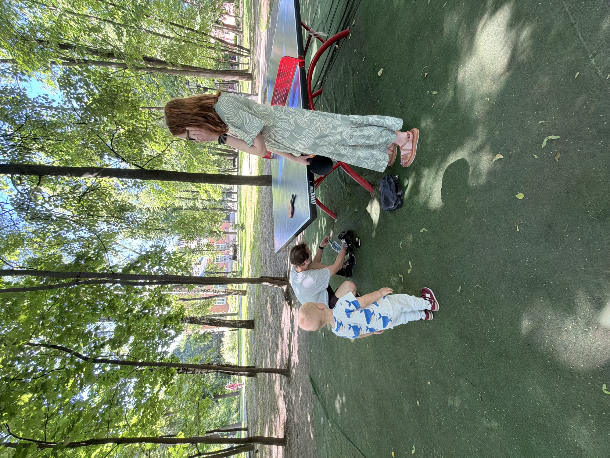 In Vidnoye, Russia, on a sun-dappled green surface in a park, a young child wearing a white shirt with blue whale patterns and grey pants stands facing a ping-pong table. A man in a light grey t-shirt and dark shorts is kneeling by the table, appearing to interact with a black object. A young woman with reddish-brown hair and glasses, wearing a light green patterned dress and brown sandals, stands near the table, holding a black ping-pong paddle and touching her face. The ping-pong table is positioned on the green surface, with trees and buildings visible in the background. A black bag rests on the ground near the woman's feet.