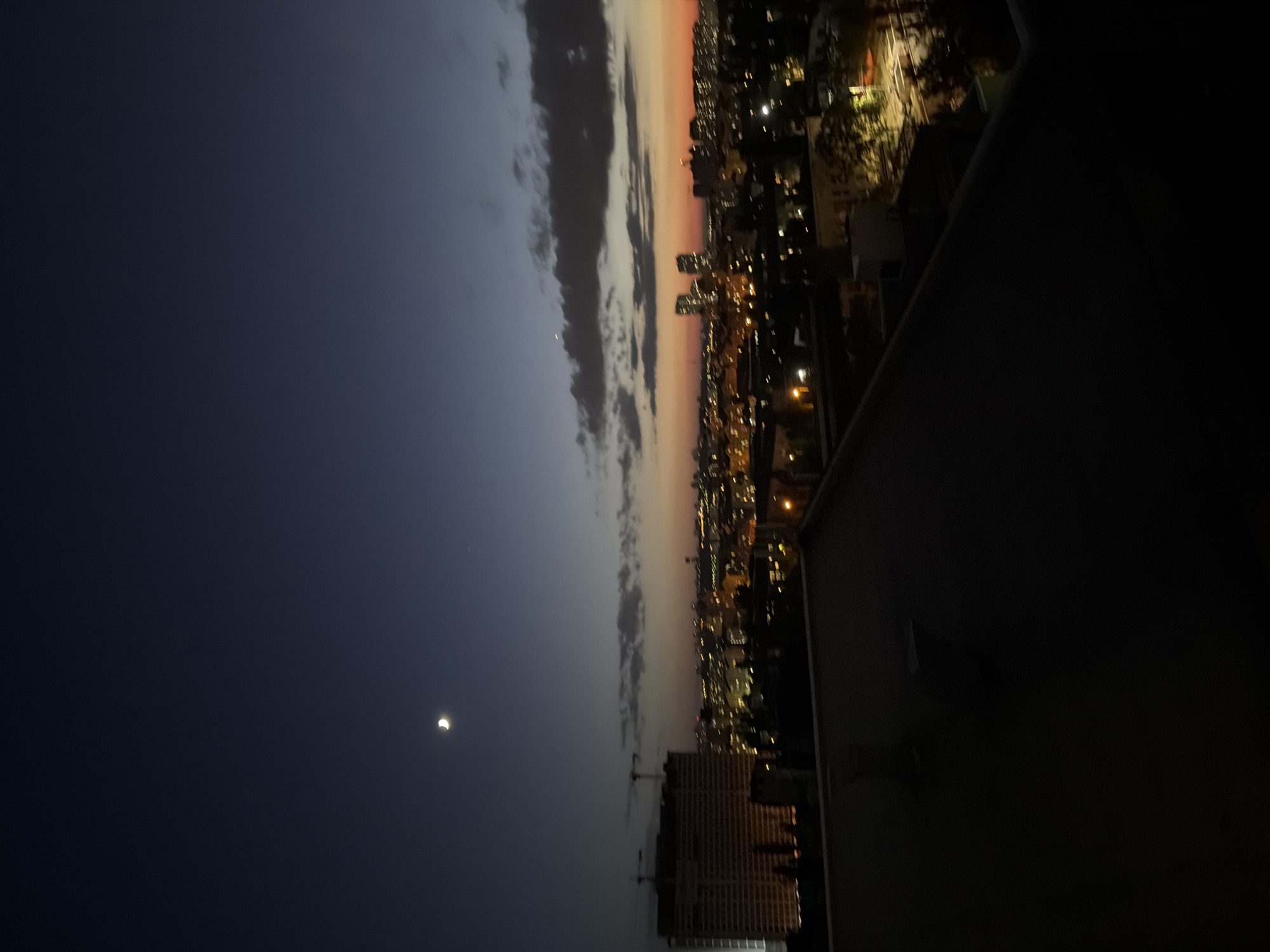 The image depicts a twilight cityscape, likely Kyiv, Ukraine, viewed from an elevated position, possibly a rooftop. The sky transitions from a deep indigo at the zenith to a warm orange glow near the horizon, indicating the period of dusk. A crescent moon is visible in the upper left quadrant of the sky. Below the sky, a sprawling urban landscape unfolds. In the mid-ground, a large, illuminated building, potentially under construction due to visible cranes, stands out on the left. Numerous other buildings with glowing windows create a tapestry of city lights. The foreground is dominated by the dark, angular surface of a rooftop, with some subtle architectural elements like vents or small structures. The overall atmosphere is one of urban tranquility at the end of the day.
