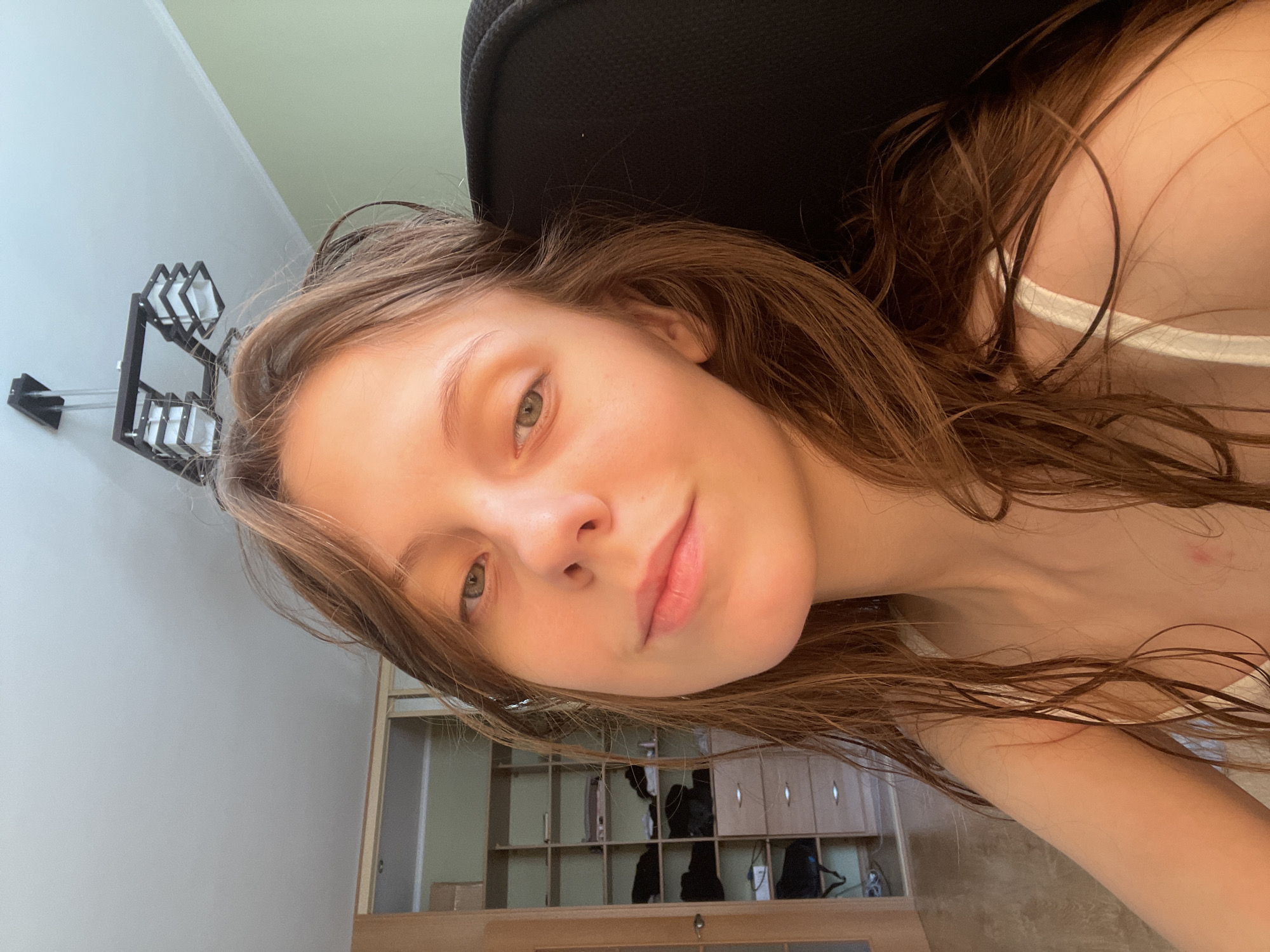 The image depicts a close-up, selfie-style shot of a young Caucasian woman with long, brown hair, appearing to be in an indoor setting. She is looking towards the camera with a neutral expression. Her skin appears to be lightly tanned, and she is wearing a white strap, suggesting a top or dress. Her hair is wet and styled loosely around her face and shoulders.
In the background, to the left, a light-colored closet or wardrobe is visible with multiple shelves and drawers, containing various items, some appearing to be clothing or personal belongings. To the right, a portion of a black chair is visible, and a light green wall is present. Above the woman's head, a modern black chandelier with multiple light fixtures hangs from a white ceiling.
The lighting in the image suggests it was taken during daylight hours, possibly near a window, as indicated by the warm tones and shadows. The overall impression is that of a casual, personal moment captured at home.
The prompt requests the location in the city of Ufa, Russia. However, there are no visual cues within the image that definitively identify the location as Ufa, Russia. The interior elements are generic and could be found in many residential locations. Therefore, it is not possible to confirm or describe the scene as being in Ufa, Russia, based solely on the visual information provided in the image.