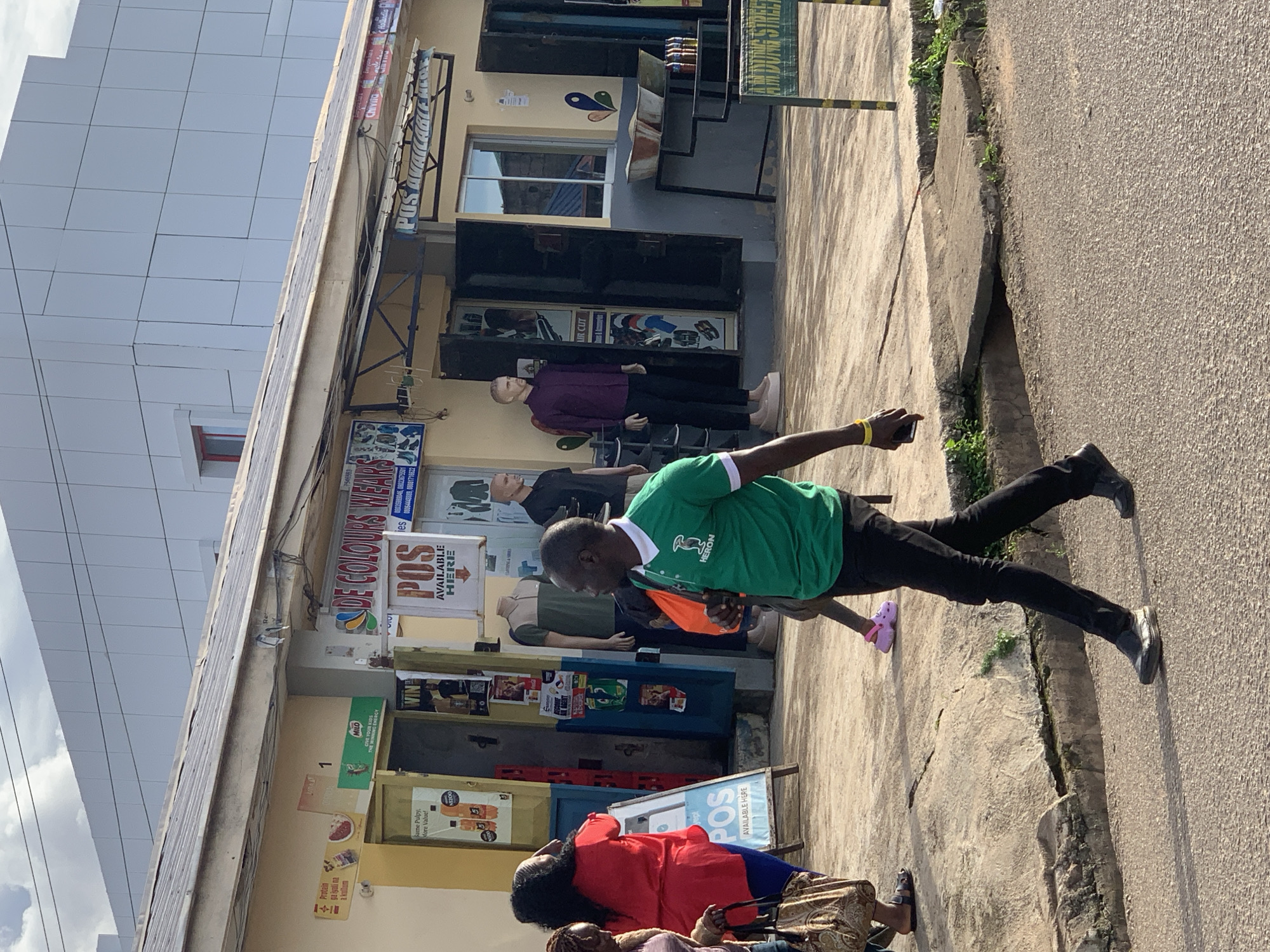 The image depicts an outdoor scene in Jos, Nigeria, focusing on a row of shops. In the foreground, a man wearing a green polo shirt with a white collar and black pants is walking. He is holding a dark-colored mobile phone in his right hand. To the left of the man, a woman in a red shirt and blue pants is visible, carrying a patterned bag. A sign that reads "POS AVAILABLE HERE" is placed on the sidewalk.
Behind the man and the woman, the shops are characterized by their open doorways and awnings. One shop has a sign that says "DE COLOURS WEARS". Mannequins dressed in clothing are displayed in the shop windows. A large, modern building with a metallic facade stands in the background, contrasting with the older storefronts. The ground is paved with concrete and asphalt. The sky appears partly cloudy.