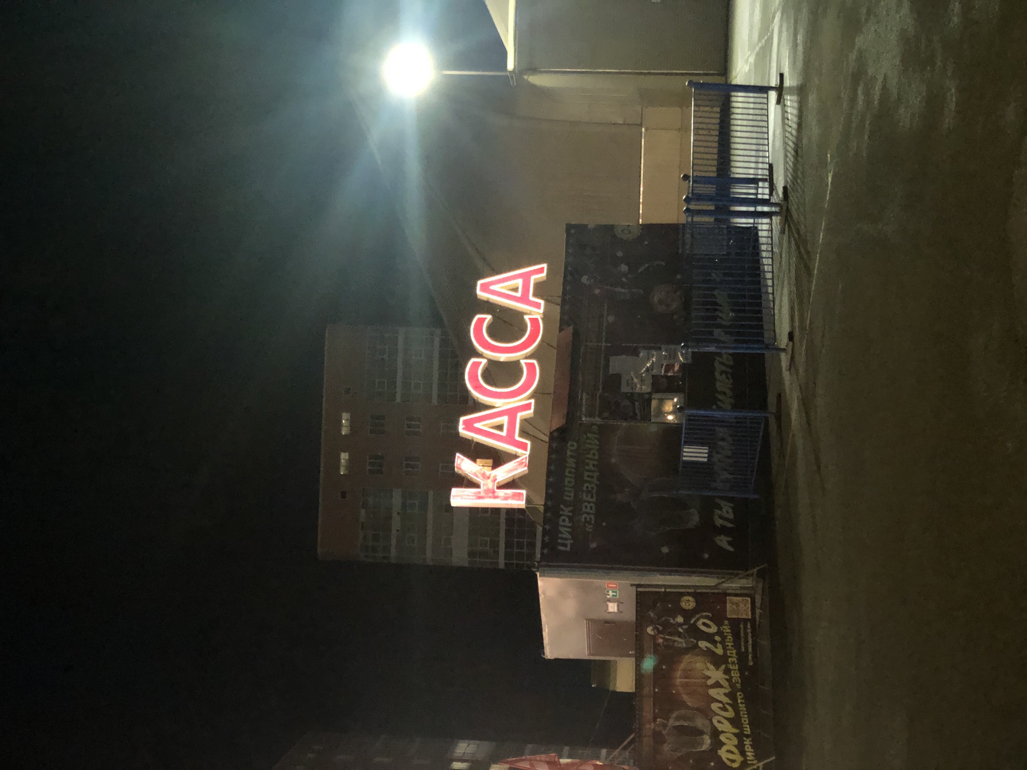 The image shows a nighttime scene in the city of Petropavlovsk-Kamchatsky, Russia. A large, bright pink neon sign spelling "КАССА" (which translates to "TICKET OFFICE" or "BOX OFFICE") is prominently displayed above a small building. This building appears to be a ticket booth or part of a venue. To the left of the booth, there is a large poster advertising "ФОРСАЖ 2.0" (which translates to "FAST & FURIOUS 2.0"), with an image of what appears to be a circus show. Below the "КАССА" sign, there is text that reads "ЦИРК шапито «ЗВЁЗДНЫЙ»" (which translates to "Circus tent 'Stellar'"). Further text below that is partially visible and difficult to decipher.
In front of the ticket booth, there are blue metal barriers, likely for crowd control. The ground is paved and appears wet or reflective, suggesting it might have rained or been recently cleaned. In the background, a tall, multi-story building with lit windows is visible. The overall lighting suggests it is dark outside, with the neon sign and a street lamp providing the primary illumination. No people are clearly visible in the image. The scene depicts the entrance area of a circus or entertainment venue.