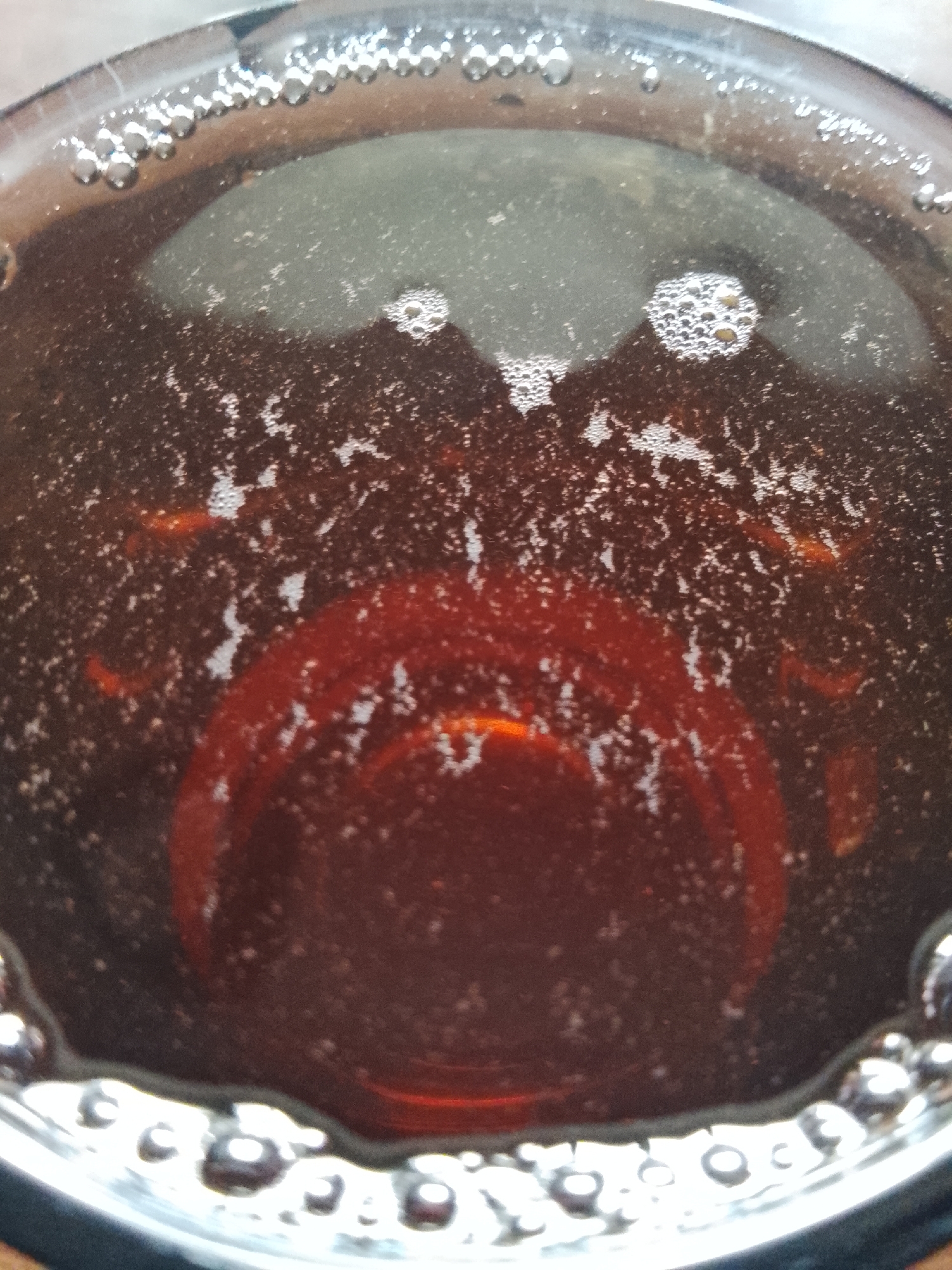 The image is a close-up, top-down view of the surface of a dark liquid in a glass. The liquid is effervescent, with numerous small bubbles scattered across the surface, particularly concentrated along the rim of the glass. The liquid itself appears to be a rich, dark brown or amber color. Reflections of light are visible on the surface of the bubbles and the liquid, suggesting it is an indoor setting or dimly lit environment. There are some larger, reddish-brown circular areas within the liquid, possibly indicating the bottom of the glass or a submerged object. No people or specific landmarks are visible. The image does not provide any information to determine its location in Almaty, Kazakhstan.