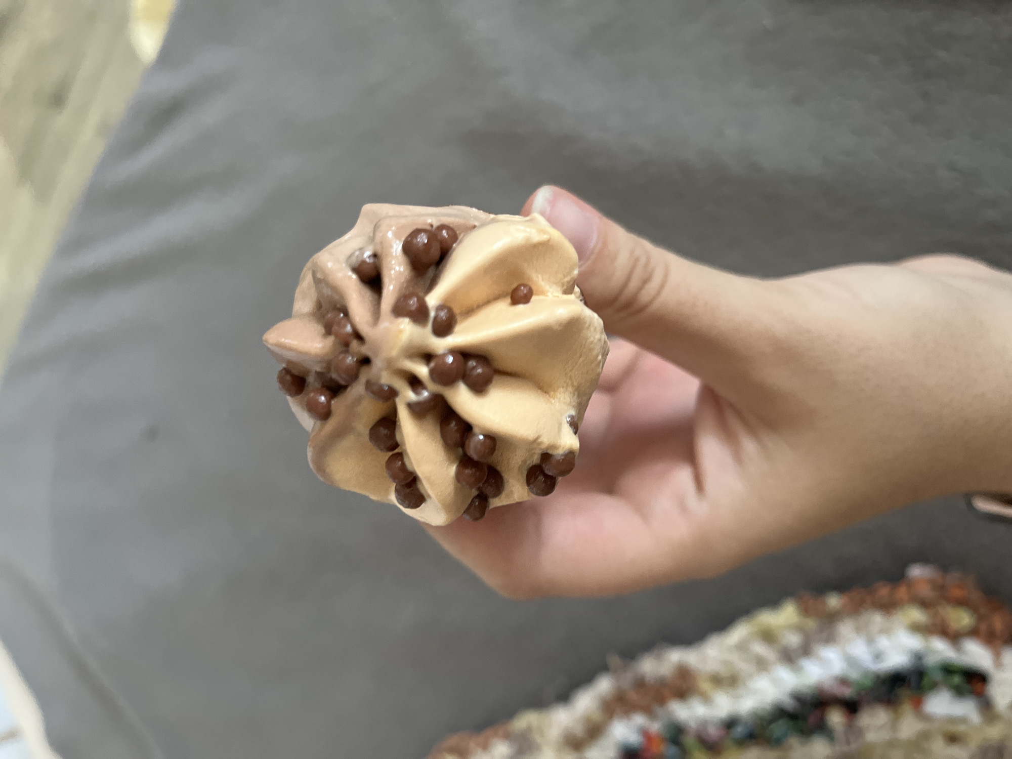 A hand holds a small, fluted ice cream cone topped with chocolate pieces. The ice cream is a swirl of two shades of brown, suggesting chocolate and caramel flavors. The background is a blurred gray surface, possibly fabric, with a colorful, textured object on the left side. The scene appears to be indoors. There is no visible indication of the location being in Krasnyye Barrikady, Russia.