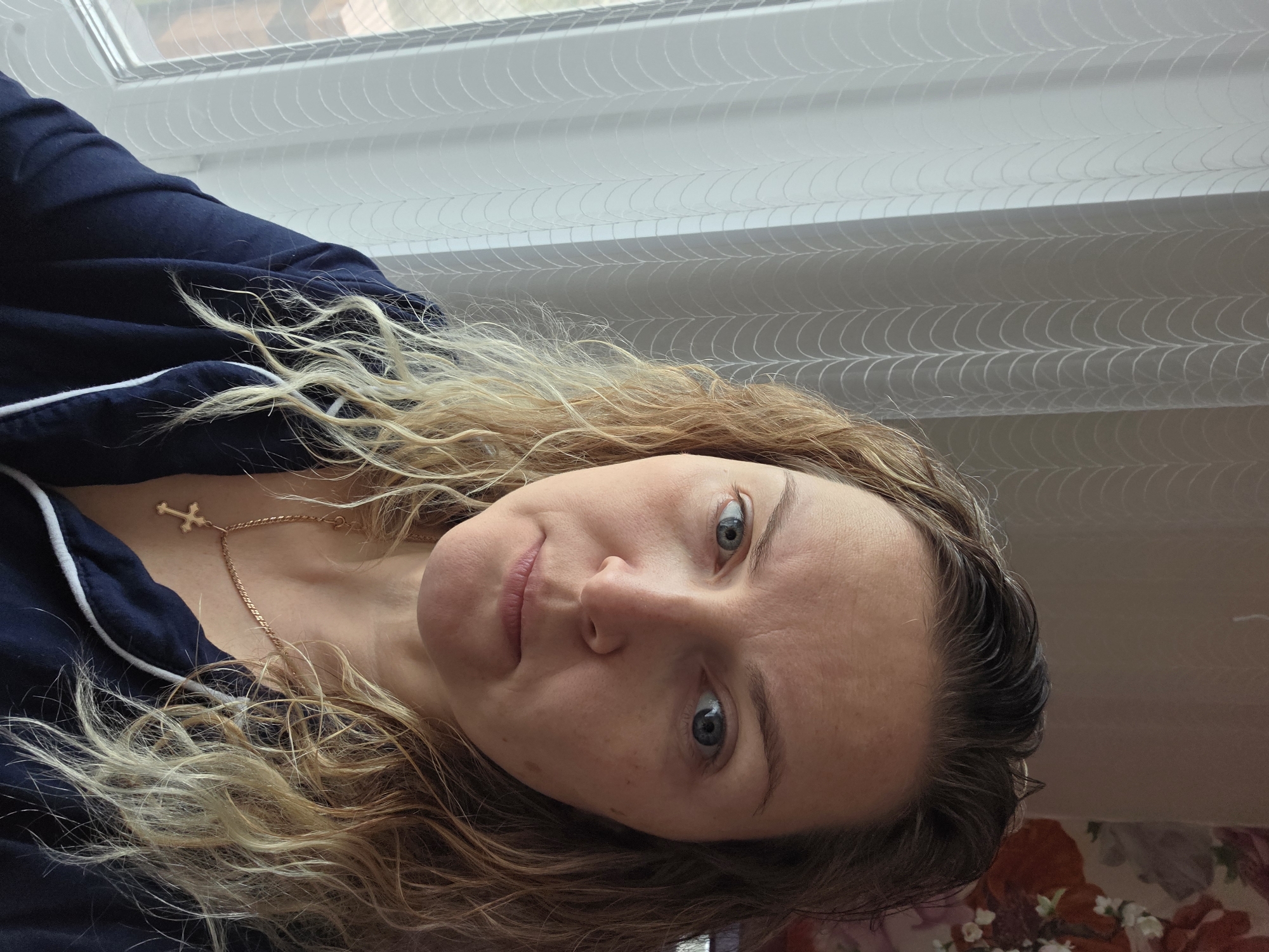 A woman with fair skin, blue eyes, and curly blonde and brown hair is facing the camera. She is wearing a dark blue top with a white trim and a gold cross necklace. The background includes a window with white blinds and a patterned surface below, possibly a table or a floor. The image was taken in a location in Chaikovsky, Russia.