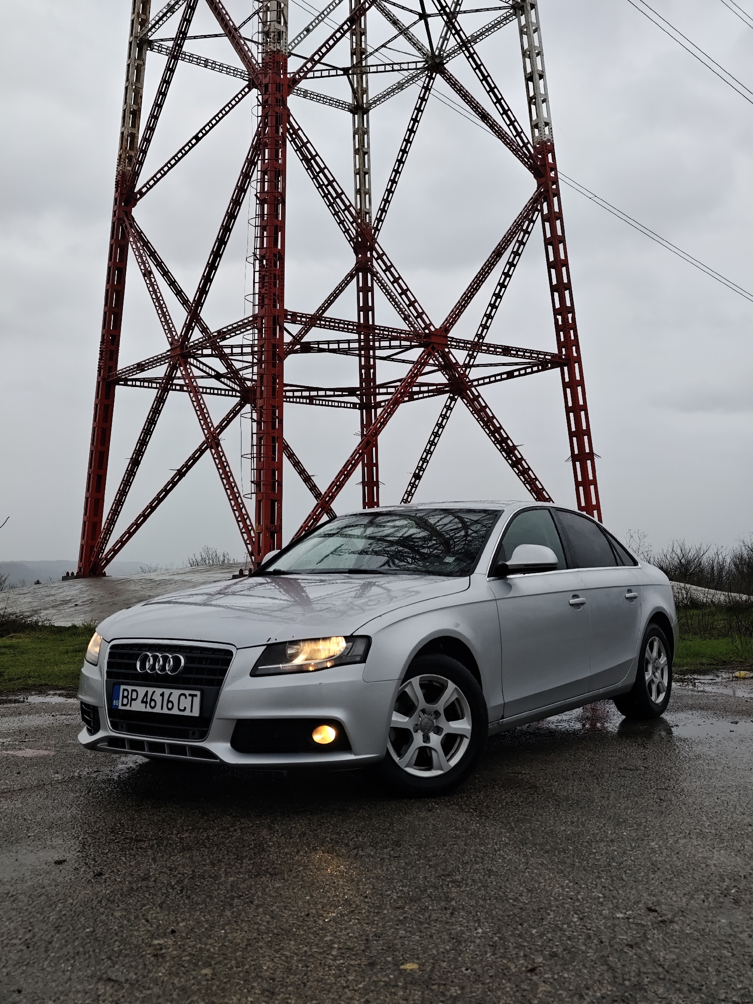 The image captures a silver four-door Audi A4 sedan parked on a wet, dark asphalt surface in Kozloduy, Bulgaria. The vehicle's headlights and left front fog light are illuminated. A Bulgarian license plate, "BG BP 4616 CT," is affixed to the front. Dominating the background is a large, weathered, lattice-style metal transmission tower, painted red in its lower sections and a lighter color higher up. Several power lines extend from the tower's upper structure. The surrounding environment consists of wet ground, patches of green grass, and sparse, dry vegetation. The sky is uniformly overcast and gray. No individuals or specific actions are depicted.