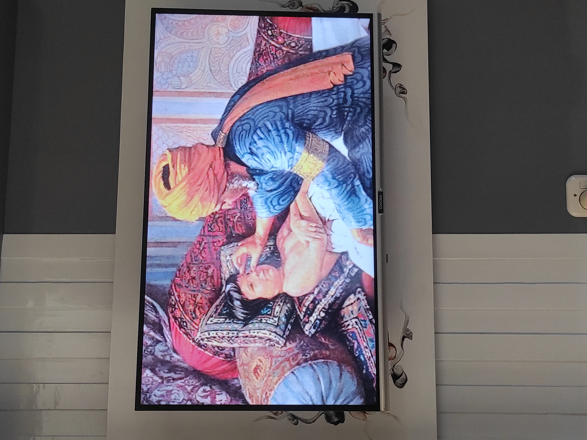 The image shows a television screen displaying a painting. The painting depicts an older person, possibly a man, wearing a yellow turban and blue robes, tending to a young child lying on ornate cushions. The older person's hand is near the child's face, suggesting an examination or caregiving action. The child is unclothed from the waist up and appears to be lying down. The background of the painting features richly patterned fabrics and architectural elements. The television is mounted on a wall in a room, with vertical blinds visible in the lower part of the frame. The image does not provide any specific information that would allow for identification of its location within the city of Sumedang, Indonesia.