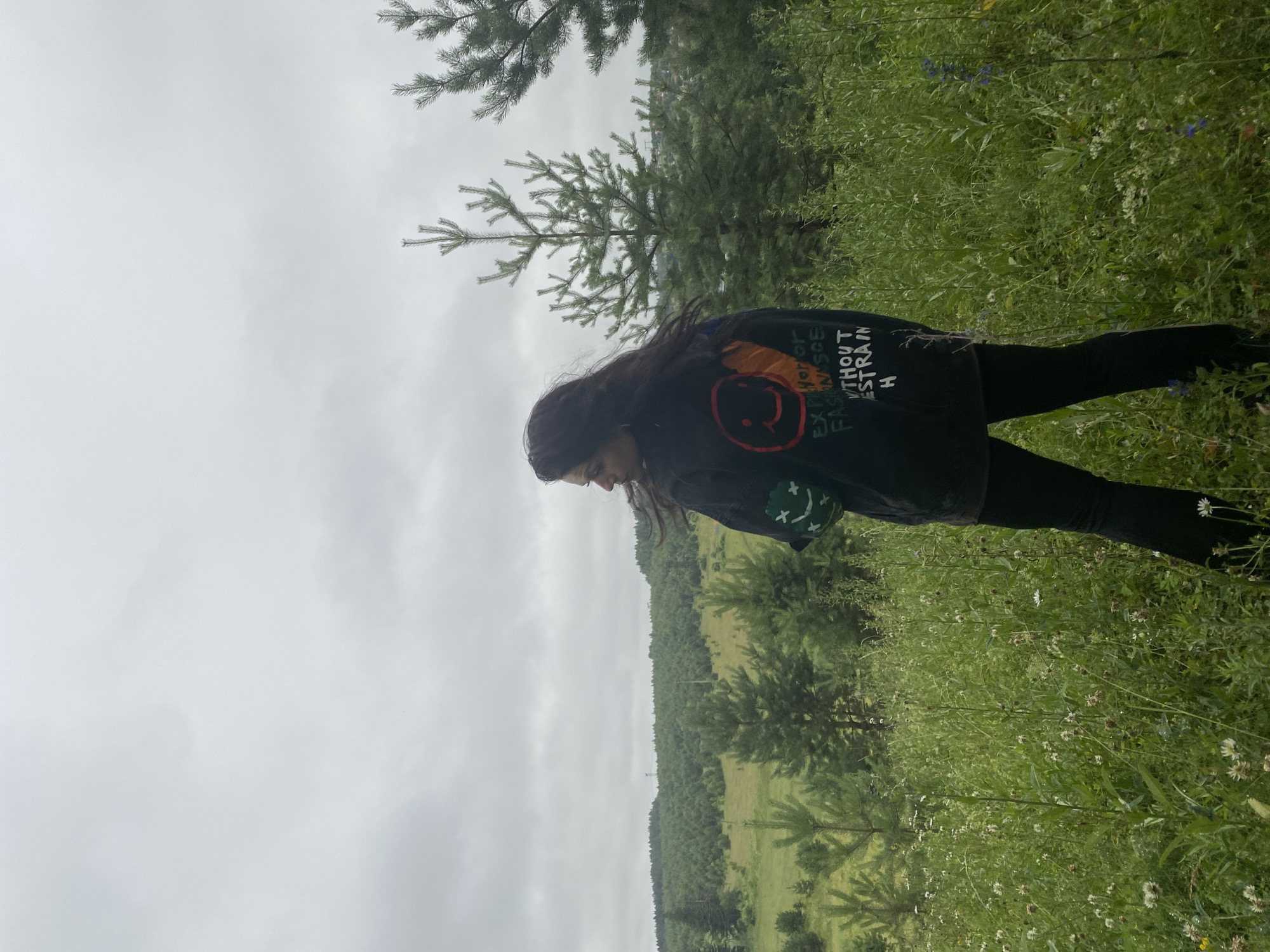 A woman with long, dark hair stands in a grassy field with scattered wildflowers and young pine trees. She is wearing a dark jacket with graphic designs on the back, including a smiley face and text. Behind her, the landscape transitions into a dense forest with rolling hills. The sky is overcast with a light gray hue. The image appears to be taken outdoors in a natural setting, likely on the outskirts of the city of Foky, Russia, given the prevalence of pine trees and the hilly terrain.