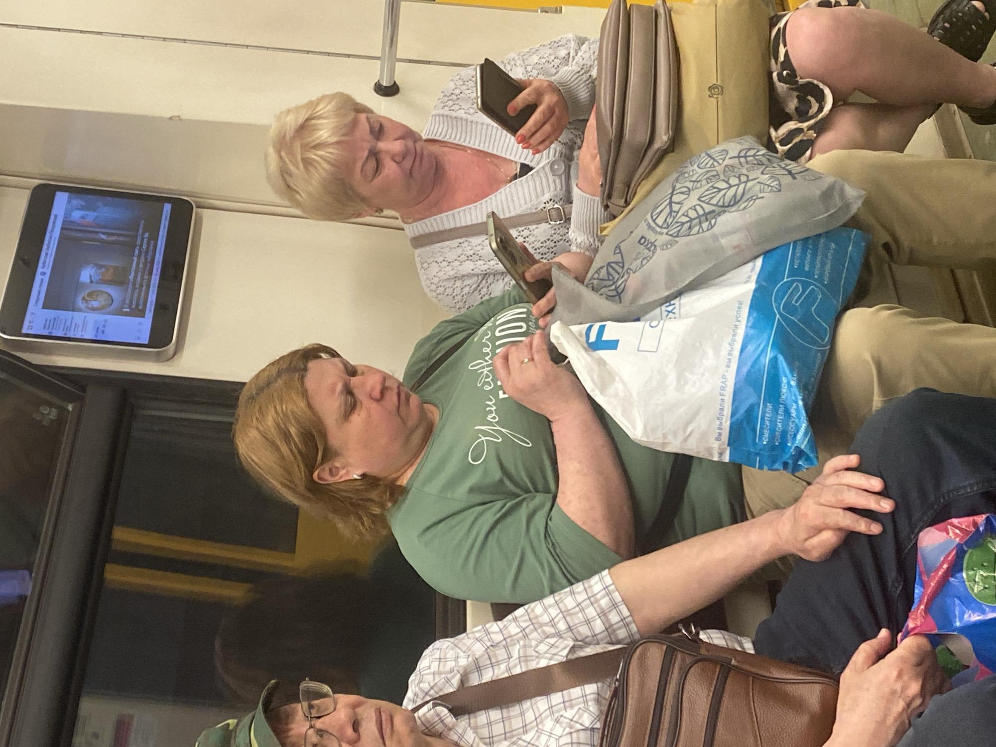 The image depicts a scene inside a Moscow, Russia metro train. Several people are seated on a bench, including two women in the upper portion of the frame. One woman, with blonde hair, is holding a phone. Another woman, wearing an olive-green shirt, is also looking at a phone. Another passenger is partially visible at the bottom of the frame, wearing a checkered shirt, glasses and a hat. Shopping bags are located on the bench. A TV screen is visible on the wall of the metro car.