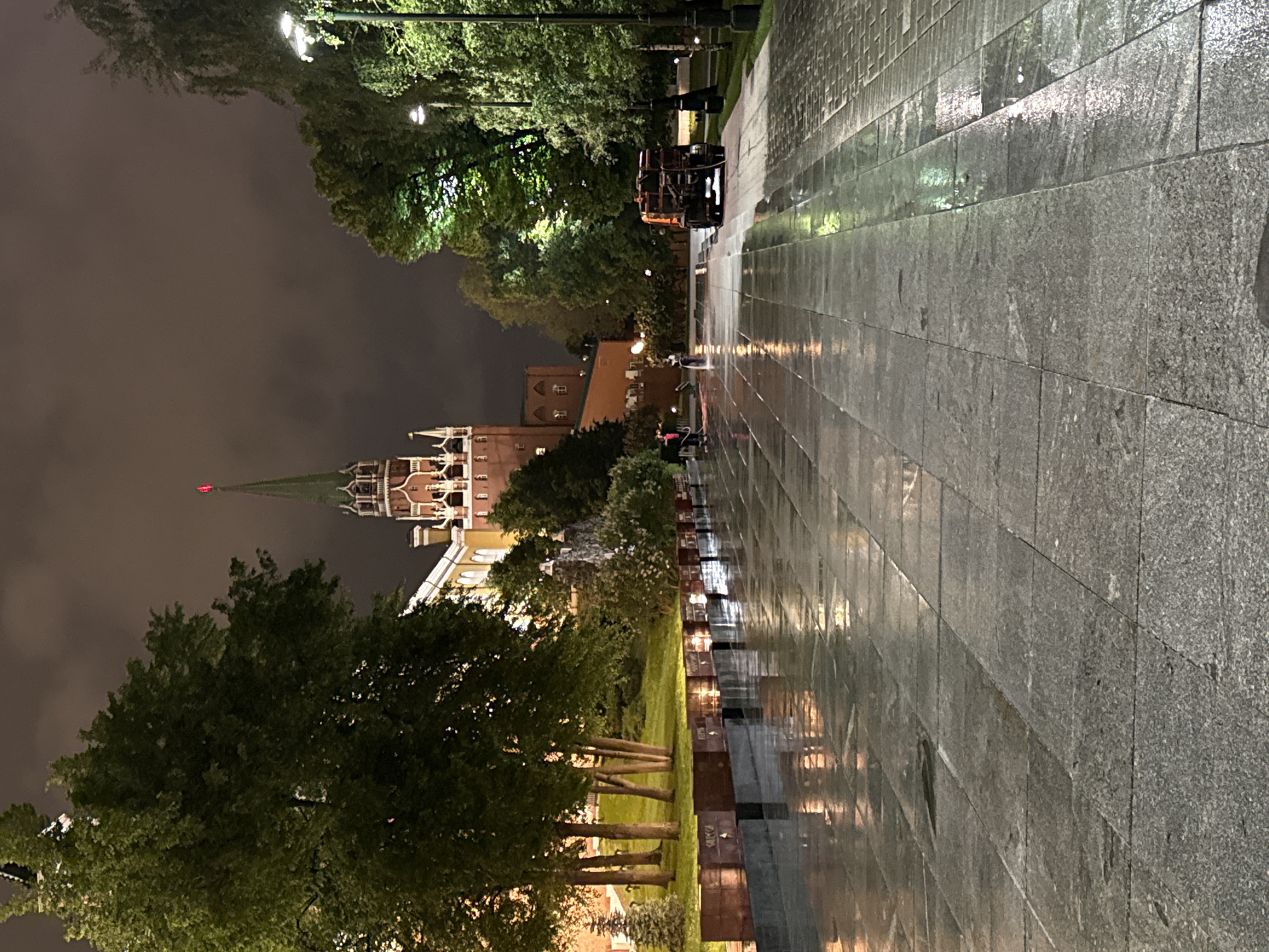 At night in an unknown location within the city of Unknown, a street with a wet stone surface is seen reflecting light. Adjacent to the street is a grass area with sparse trees and markers. In the background is a structure with a notable pointed tower, possibly a part of a castle or fortress, and a dark, cloudy sky. A vehicle is visible further down the street, under lit street lamps.