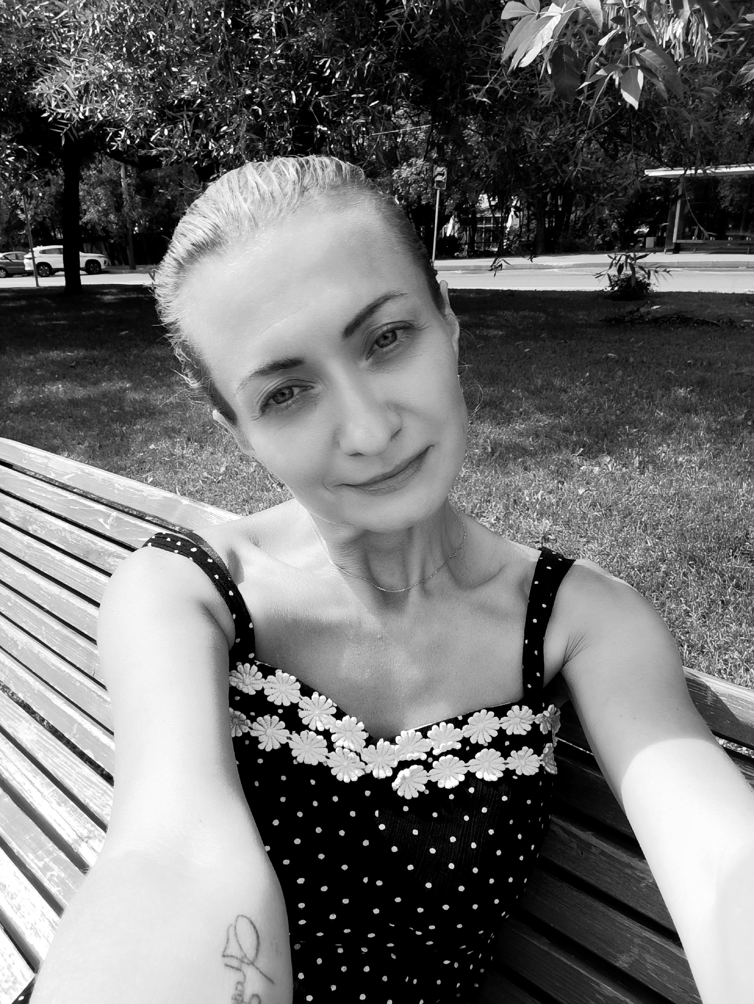 A black and white, head-and-shoulders selfie of a woman taken outdoors. The woman is sitting on a slatted wooden bench, facing the camera. She has blonde hair pulled back, and is wearing a dark, polka-dotted dress with white floral embellishments along the neckline. Her left arm is visible, resting on her lap and showing a tattoo on her forearm. The background consists of a grassy area with trees, and a paved path with a bus stop shelter and vehicles further in the distance. The lighting suggests it is daytime, with shadows cast on the grass. The location is identified as Moscow, Russia.