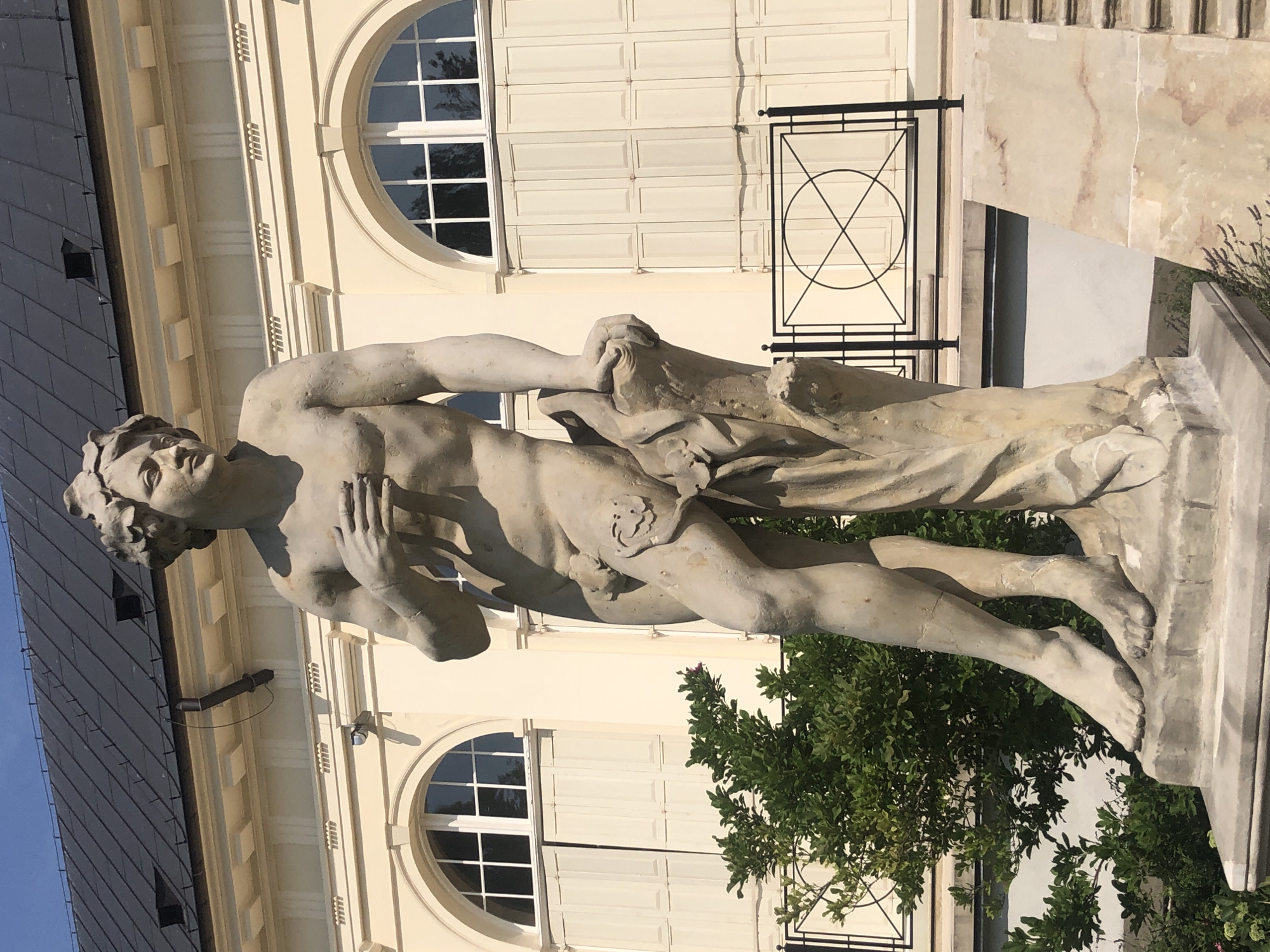 The image shows a male statue in front of a building. The statue is made of light-colored stone and depicts a nude man with curly hair. He is standing with his left leg slightly forward, and his right arm is resting on a tree trunk behind him. His left hand is placed over his chest. The building behind the statue is light yellow with large arched windows and white shutters. A black metal fence with a circular design is visible in front of the building. Green bushes are planted to the left of the statue. The sky is blue. This statue is located in the Wilanów Palace complex in Warsaw, Poland.