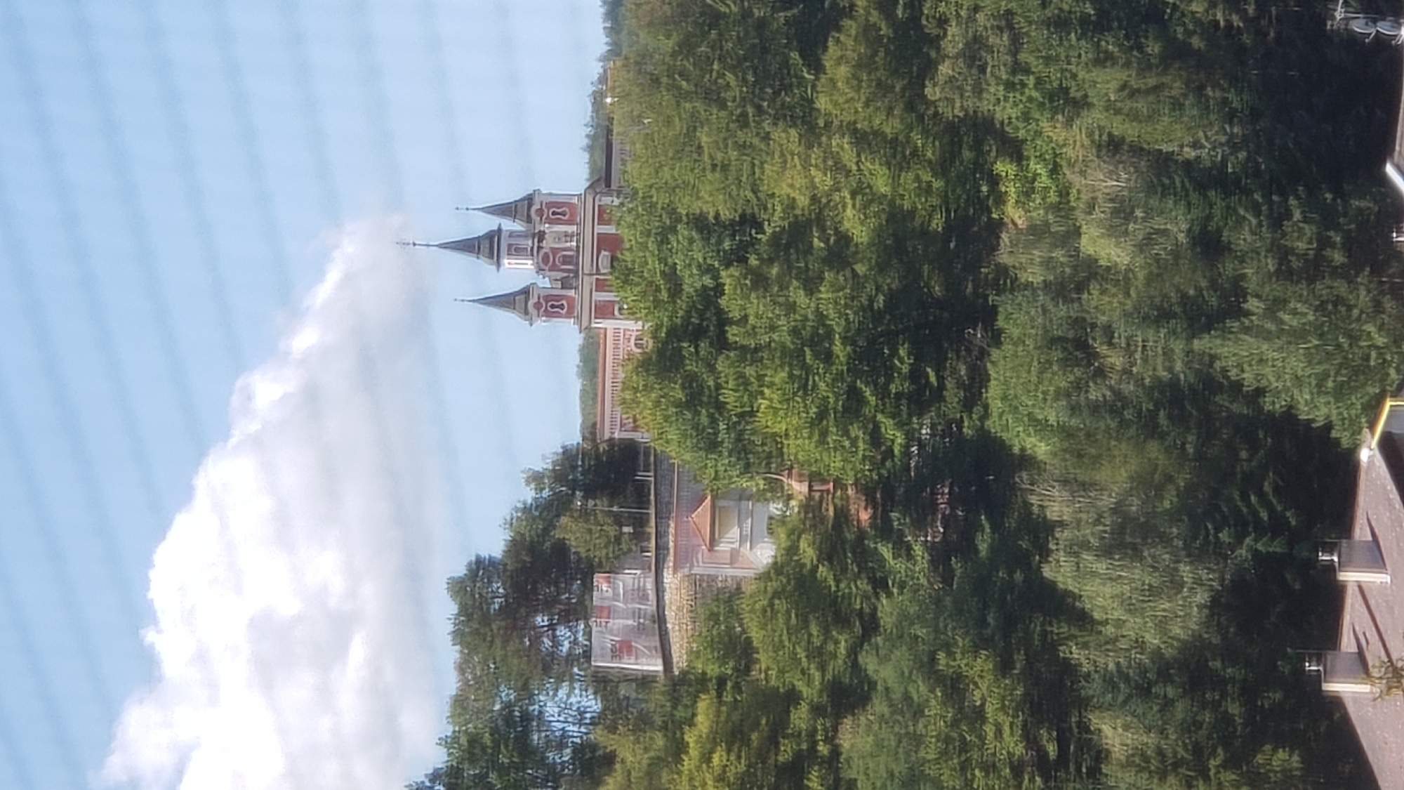 The image shows a wide shot of a historical church with red and white accents and multiple spires, perched on a hill amidst lush green trees. The church appears to be part of a larger complex, with some scaffolding and white coverings visible on lower structures to the left. In the foreground, the roof of a building with two chimneys can be seen. The sky above is blue with some scattered white clouds, and horizontal lines are visible, suggesting the photo was taken through a window with blinds. The scene is set in a hilly, forested environment, likely a semi-urban or rural area. This specific church is the Orthodox Cathedral of St. John the Merciful in Prešov, Slovakia.