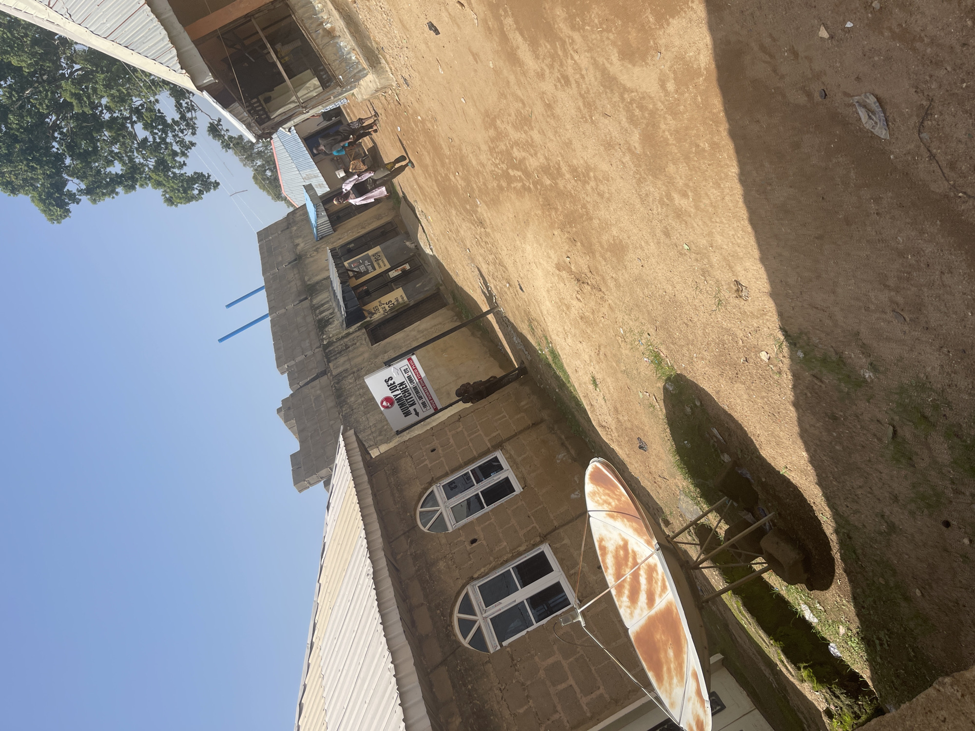 In Jos, Nigeria, a satellite dish, showing signs of rust and wear, sits on a metal stand in front of a beige brick building with arched windows. A sign for "Mummy Joe's Kitchen" is visible on the building's exterior. The ground is a mix of dirt and sparse grass. In the background, several other buildings with corrugated metal roofs are present, and a few individuals are walking on a dirt path. The sky is clear and blue.