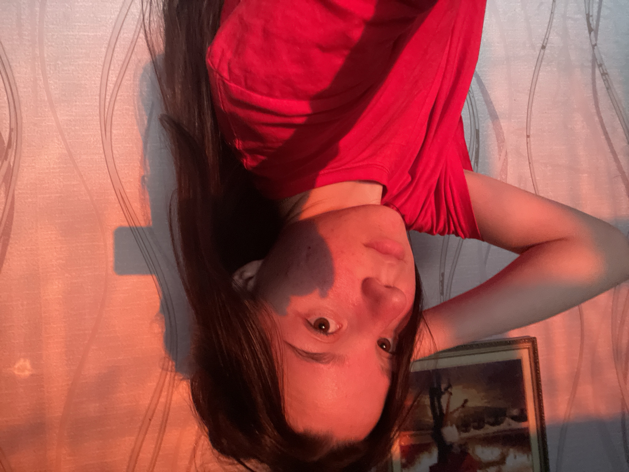 A young woman with long dark hair, wearing a red t-shirt, is captured in a selfie against a textured wall. The scene is illuminated by warm, orange light, casting shadows that create an abstract pattern on the wall and on her face. Her left arm is raised, with her hand behind her head. A framed picture hangs on the wall to the left of the subject. The environment appears to be indoors, possibly a room in a residential building. No specific details about the city of Olkhovka, Russia, can be inferred from the image itself.