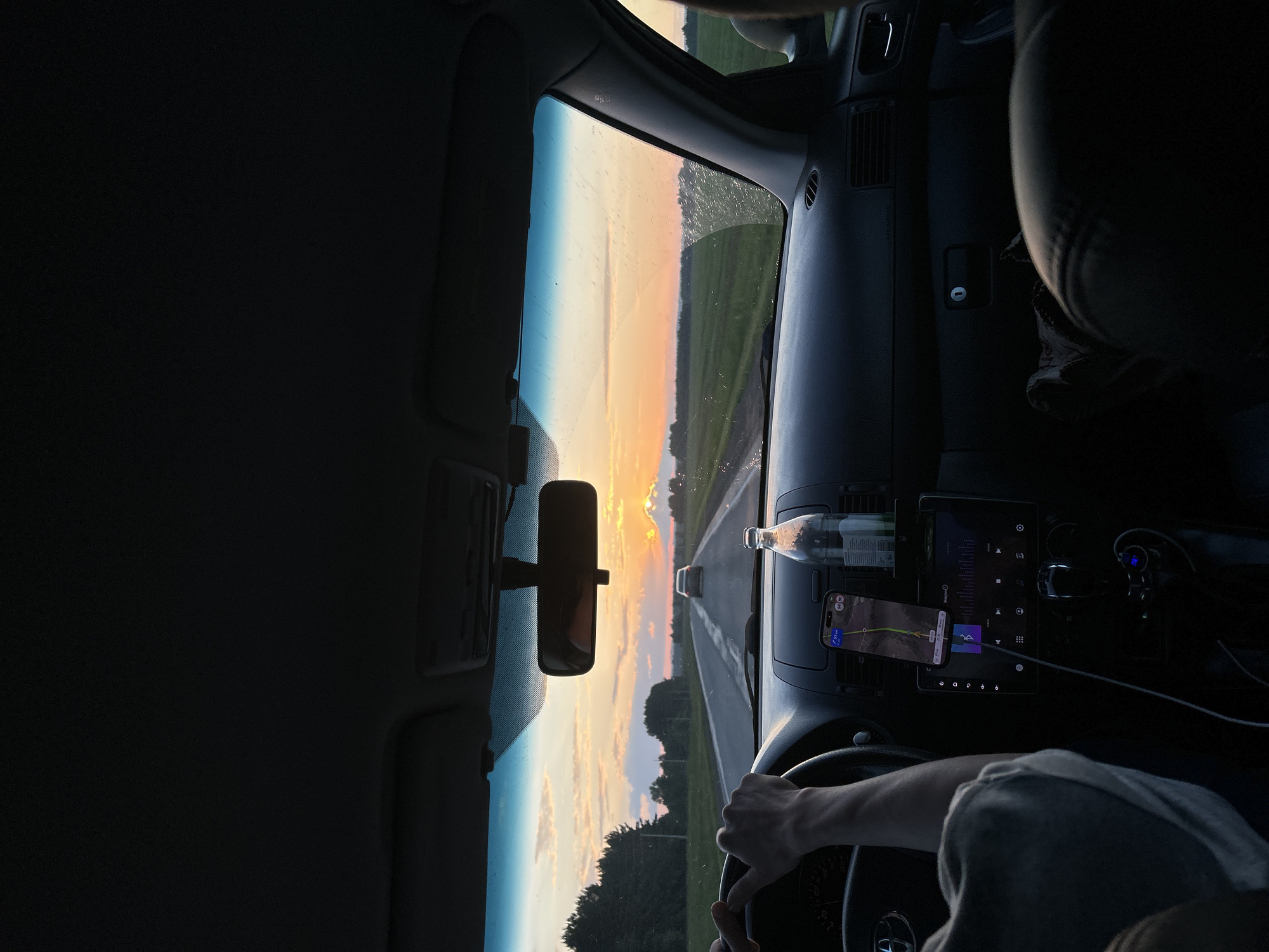 A first-person perspective shot from inside a car driving on a rural road in the city of Irnyshki, Russia, at sunset. The sun is setting on the horizon, casting a warm glow across the sky with shades of orange and yellow. The road ahead is straight and empty, flanked by green fields and trees. The car's dashboard is visible, with a phone mounted on a holder displaying a navigation app. A bottle of water is also placed on the dashboard. The steering wheel and the driver's hands are in the foreground, indicating that the car is in motion. The overall mood of the image is peaceful and serene, capturing the essence of a scenic drive during golden hour.