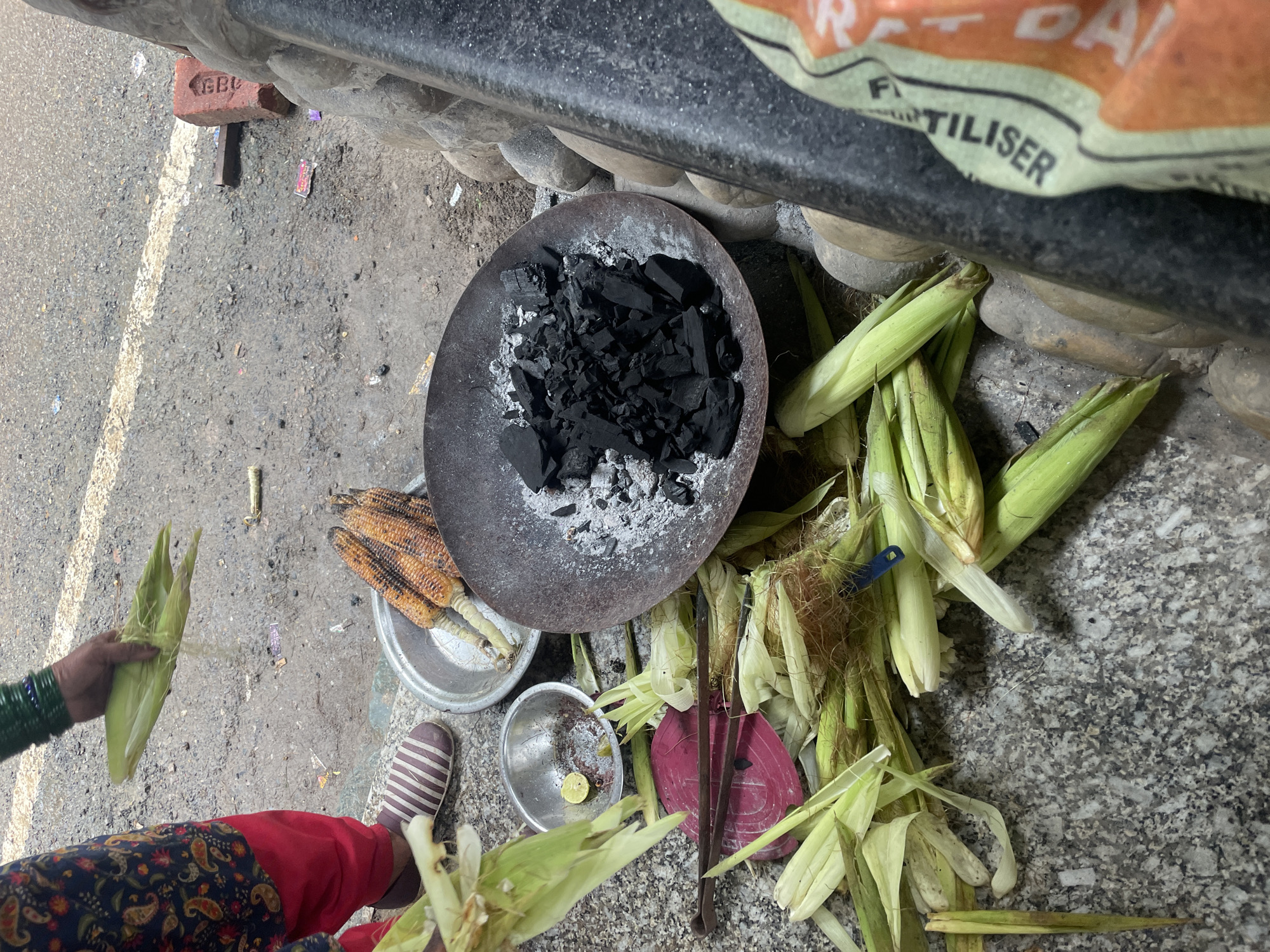 In Bhagsu, India, at ground level, an individual's hands are seen holding a husk of corn, preparing it for roasting. A collection of corn cobs, some roasted and others raw with husks, are arranged on the ground. A metal bowl containing charcoal sits nearby, indicating the roasting process. Another small metal bowl holds a piece of lemon and some remnants of spices. The scene unfolds on a gritty surface, with a prominent stone wall to the right. The overall impression is one of an outdoor food preparation setting.