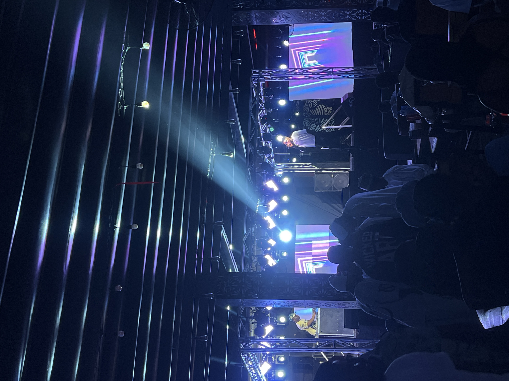 The image depicts a live performance in what appears to be a nightclub or concert venue. The venue is characterized by vertical, corrugated wall panels and spotlights, and a stage with multiple levels of lighting and video displays. Several people are visible in the audience and on the stage. A performer is stationed on one of the platforms, and audience members are positioned around the front of the stage. The overall atmosphere is one of a live event in a dark, illuminated setting. Given that the location is Howayi, Nigeria, it is likely that this is a modern entertainment venue in a bustling urban area.