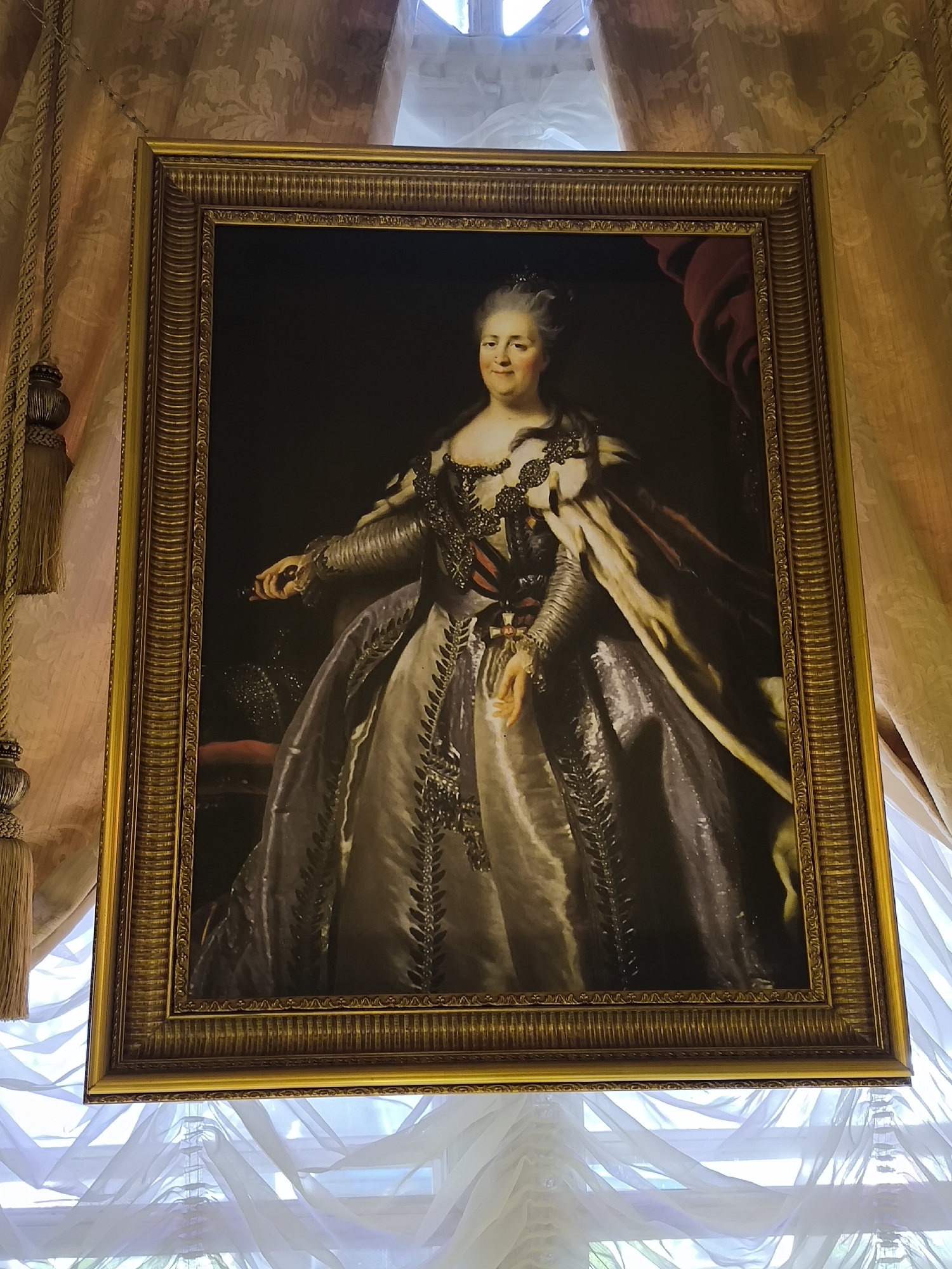 This is a portrait of Catherine the Great, Empress of Russia, painted in Saint Petersburg. She is depicted in a formal, regal pose, wearing an elaborate gown with a fur-lined cloak draped over her shoulders. Her attire is adorned with intricate embroidery and jewelry, signifying her status. She wears a small crown or tiara in her hair, and her expression is composed and serene, suggesting confidence and authority. Her left hand is slightly extended, holding an object that is not clearly discernible.
The portrait is set within an ornate golden frame, and the painting itself is hung against a backdrop of opulent drapery. To the left and right of the painting, heavy, gold-toned curtains with tassels are visible, hinting at a grand interior. The lighting appears to be from an unseen source, likely natural light filtering through windows, as indicated by the translucent white fabric draped across the bottom of the frame, which resembles sheer window coverings. The overall impression is one of wealth, power, and historical significance, consistent with the setting of a royal palace or museum in Saint Petersburg. There is no visible text in the image.