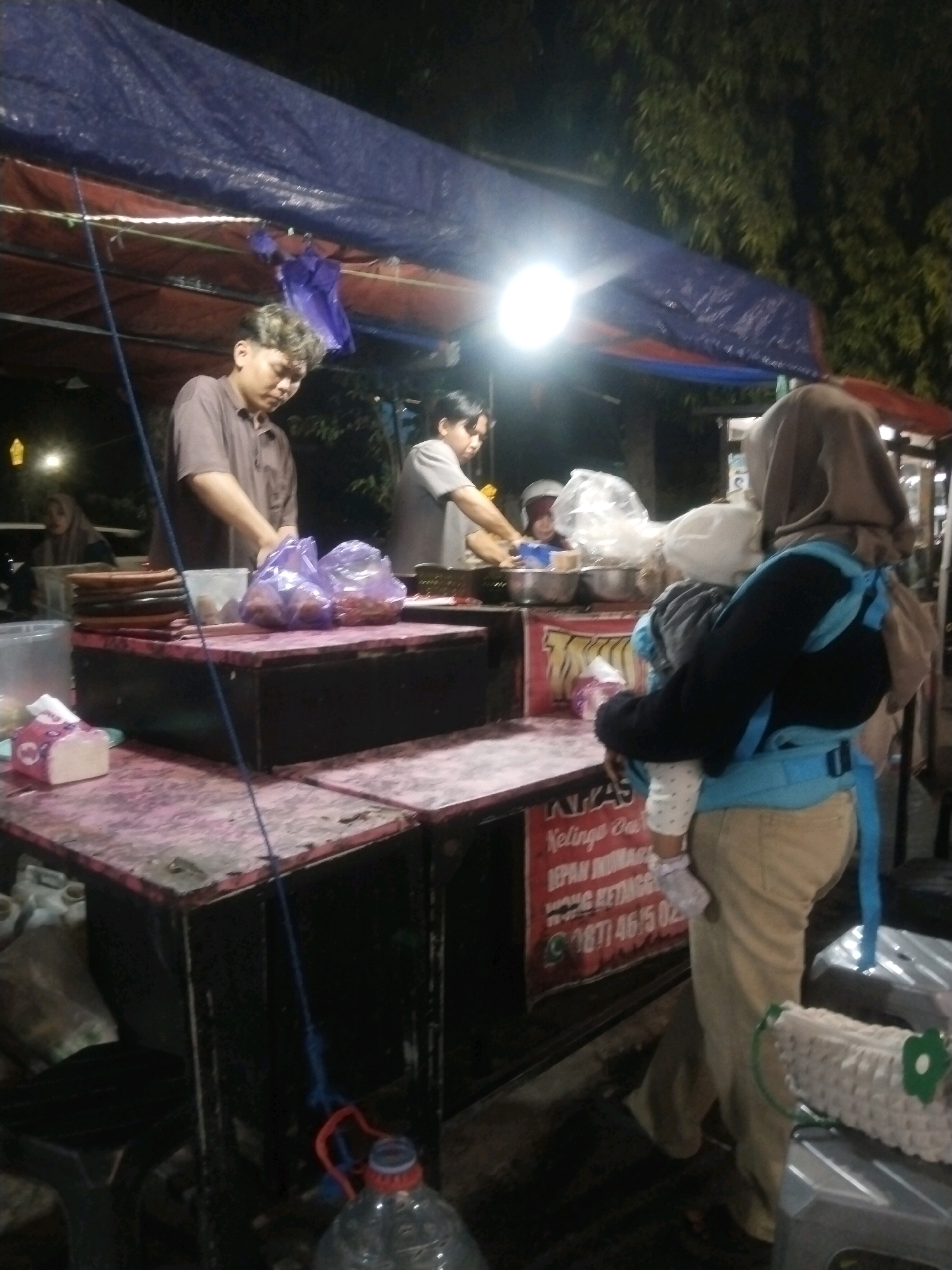 The image depicts a street food stall at night in Pemalang, Indonesia. A person wearing a light brown collared shirt is standing behind the counter, appearing to be preparing food. To their right, another person in a grey t-shirt is also working at the stall. In the foreground, a woman wearing a beige hijab and pants carries a baby in a blue front carrier. The stall is covered by a dark blue tarp, and a bright light illuminates the scene. Various food items are visible on the counter, including bags of what appear to be snacks or ingredients, and a stack of plates. A partially visible sign with Indonesian text is mounted on the stall. The surrounding environment is dark, suggesting an outdoor urban setting.