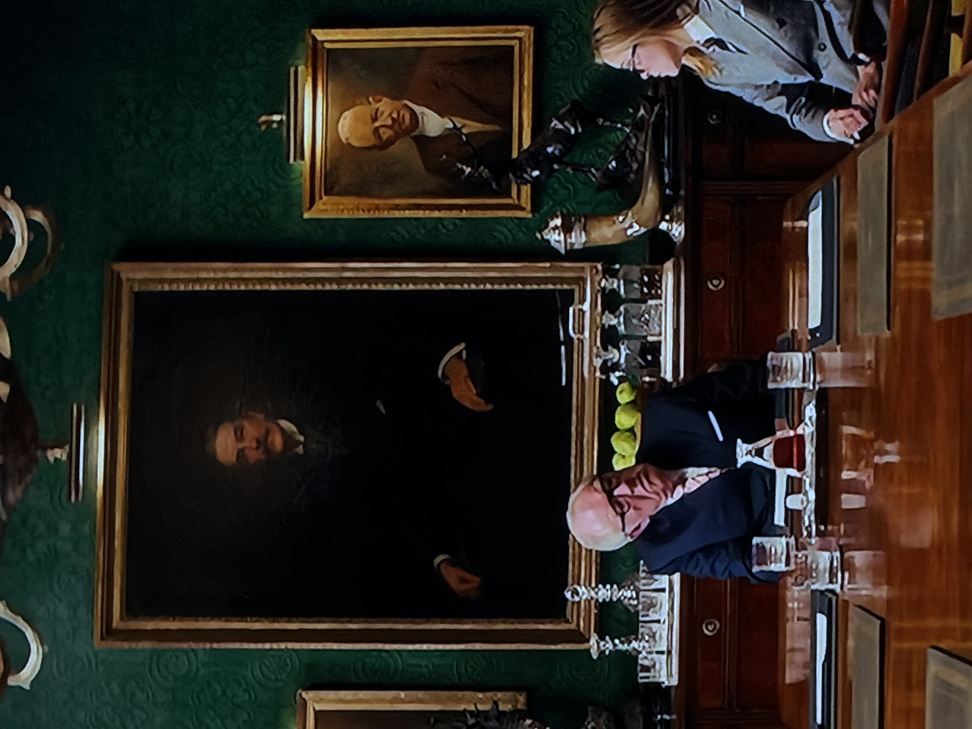 An interior shot depicts a formal room with dark green, patterned wallpaper. Two framed oil paintings of men hang on the wall, lit by sconces. The upper painting shows an older man with light hair and a white collar, while the larger lower painting features a man with dark hair and a mustache.
A long, polished wooden table occupies the right side of the frame. An older man with light skin, a balding head, and glasses is seated at the table, wearing a dark suit jacket and a light-colored shirt with a tie. His hands rest on the tabletop. In front of him are several clear glasses and a small bowl of green fruit, possibly apples or limes. Further down the table, more glasses are visible.
A younger woman with light hair and glasses stands behind the table on the left, wearing a light-colored jacket over a white top. She appears to be looking down at something on the table or an unseen element. Dark wooden furniture, possibly shelving or cabinets, is reflected in the table's surface. A dark wooden door with a round handle is visible in the background. The scene suggests a formal gathering or meal in a traditional setting in Sofiivska Borshchahivka, Ukraine. No visible text is present.
