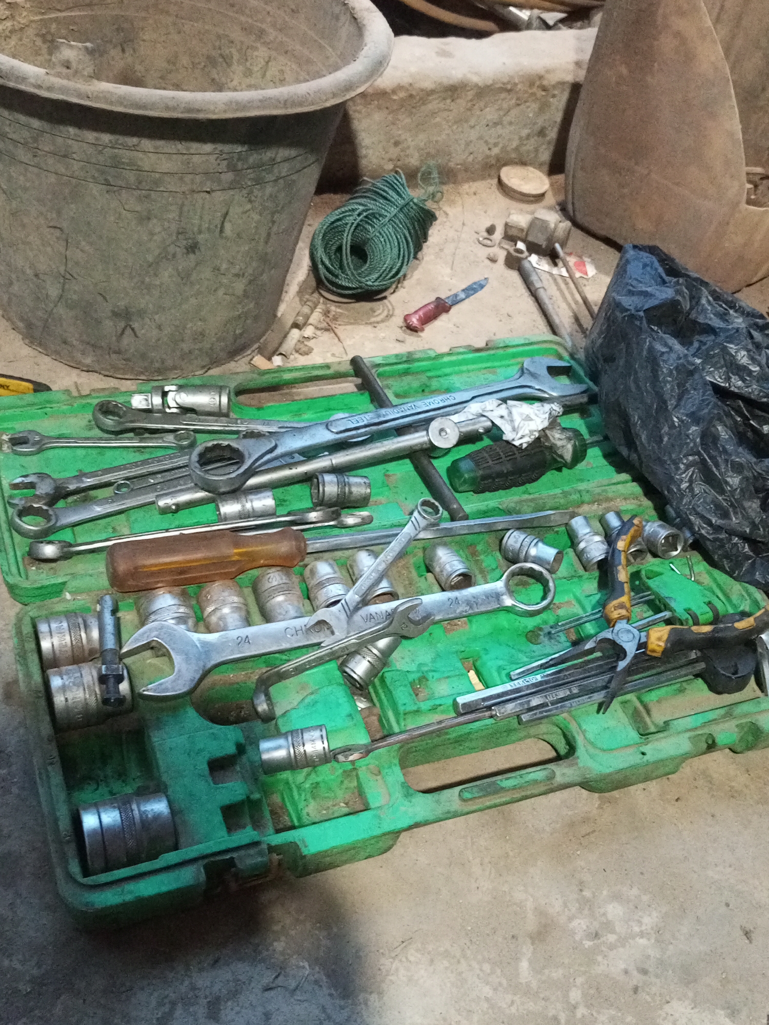 In Keling Panggau, Indonesia, a collection of tools is laid out on a concrete surface. The primary subject is a green plastic toolbox, open and filled with various wrenches, sockets, and a screwdriver with a brown handle. A length of green rope is coiled to the left of the toolbox, near a large, dark grey plastic bucket. Other items visible include a black plastic bag, several metal mallets, and a small red-handled knife. The overall scene suggests a work or storage area, possibly outdoors or in a garage.