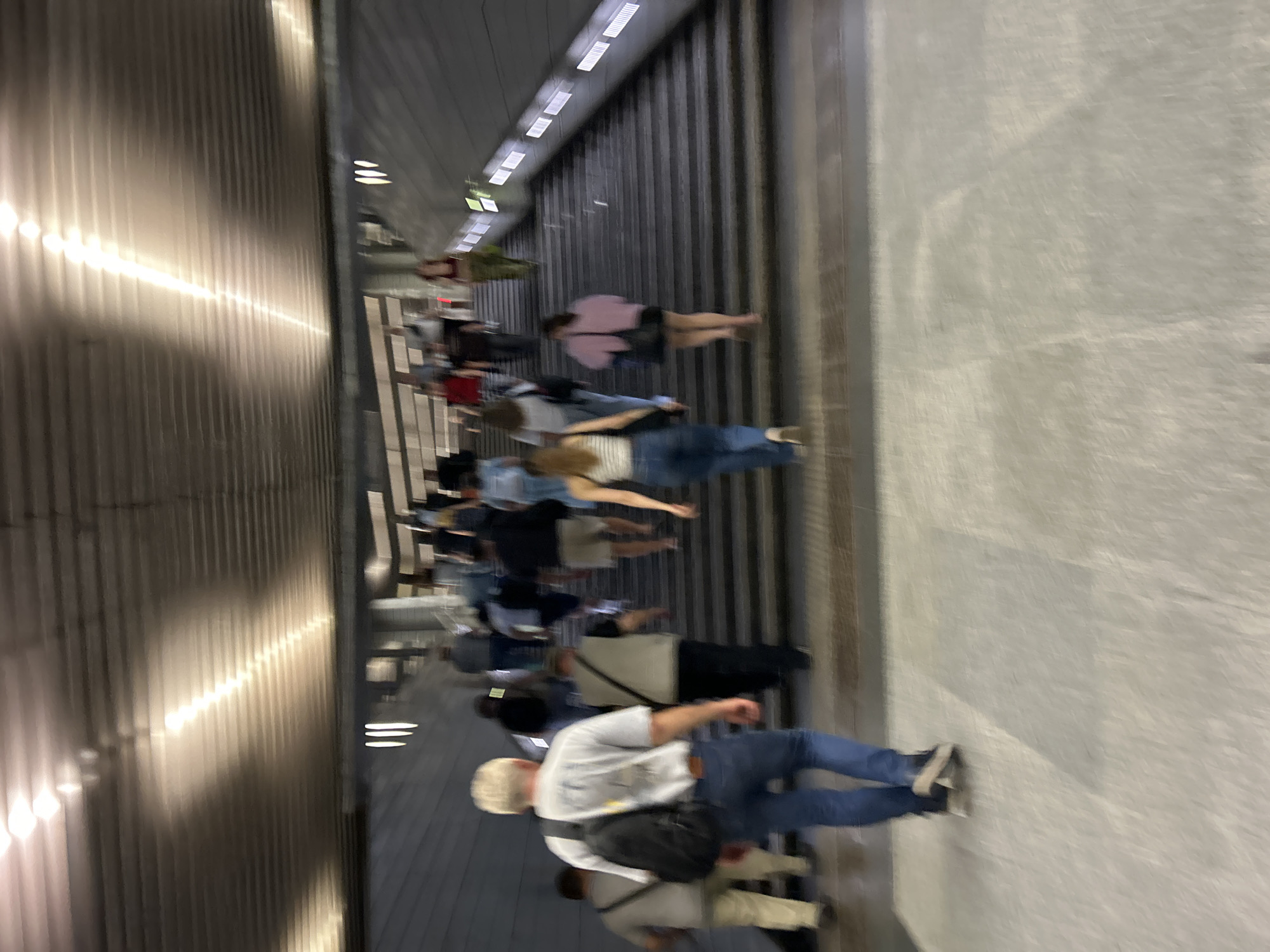 The image depicts a group of people ascending a wide, outdoor staircase in a city setting. The lighting suggests it is either daytime with artificial illumination or nighttime. The architecture features a corrugated metal ceiling with linear light fixtures. The stairs themselves are dark gray. The people are varied in their attire, with some wearing casual clothing like t-shirts, jeans, and shorts. The motion blur indicates movement and possibly a slightly unsteady camera or hurried pace of the subjects. One man in the foreground, facing away from the camera, wears a light-colored t-shirt, blue jeans, and a black backpack. Another individual, a woman in a white and black striped top and blue jeans, is also ascending the stairs. The environment appears to be a public thoroughfare or an access point to a higher level. The specific location of Moscow, Russia cannot be determined from the visual information provided.